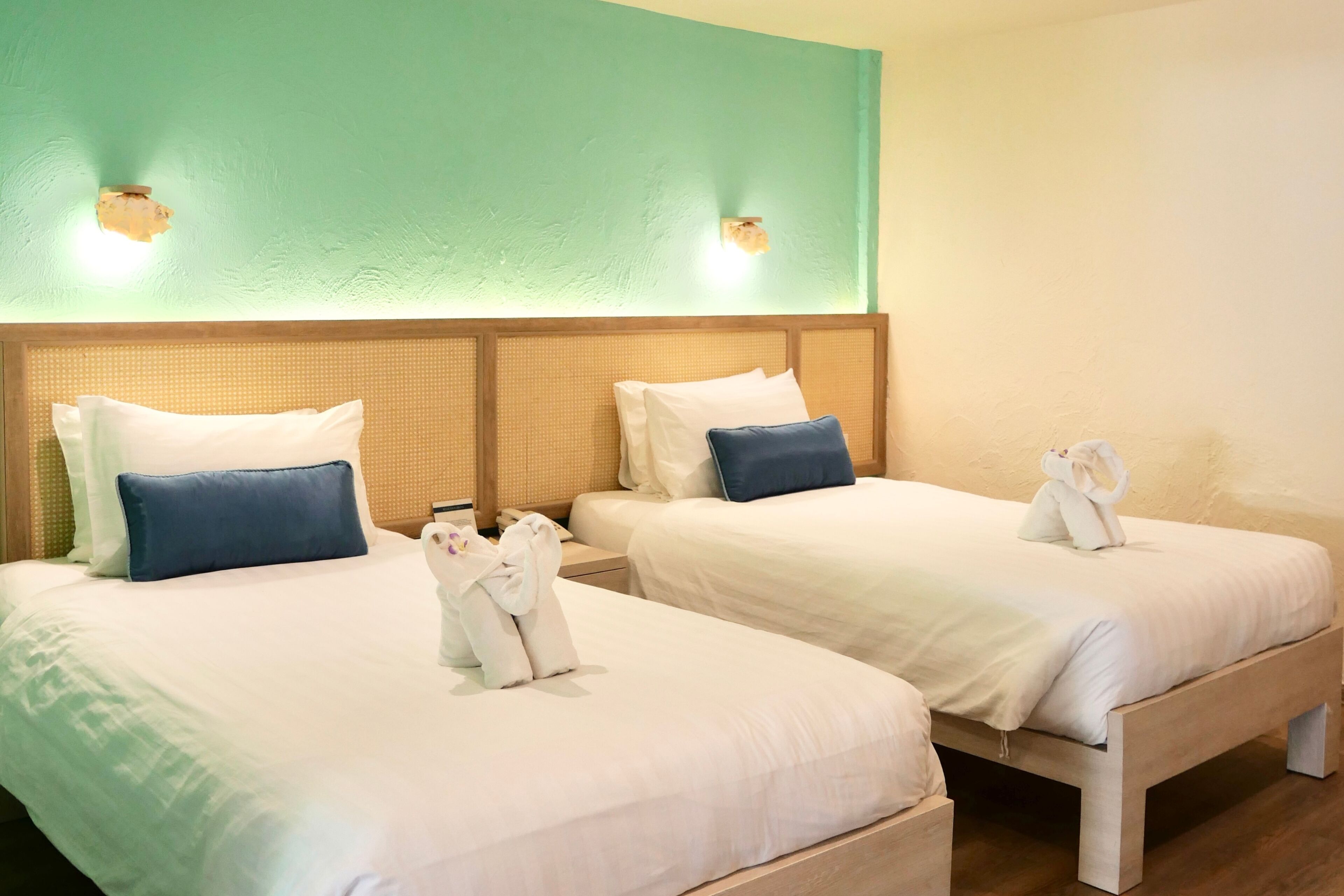 A hotel room with two beds, each with a pillow, a bedside table, and a towel rack with towels.