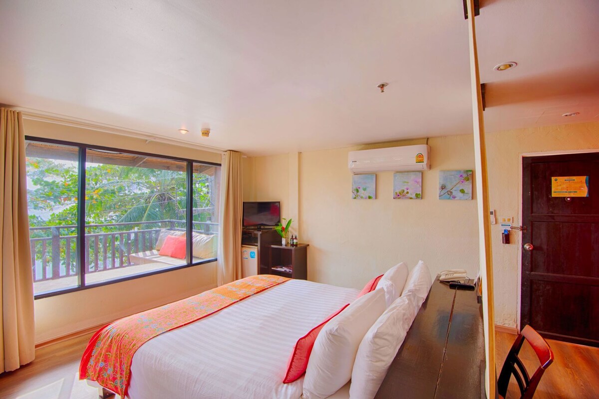 A hotel room with a large bed, a TV, an air conditioning unit, and a balcony with a view of greenery.