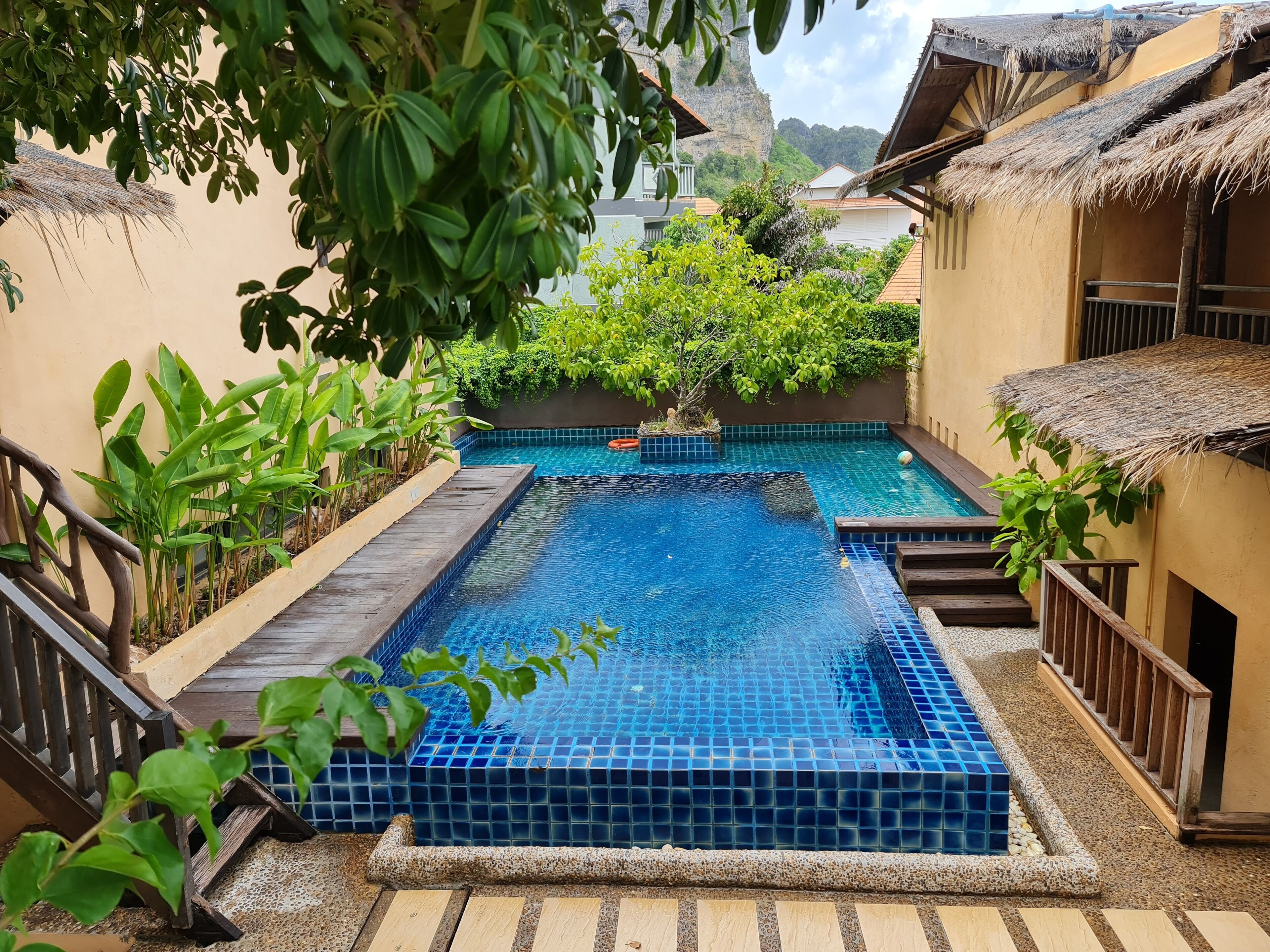 Piscina con azulejos azules, rodeada de plantas y una terraza de madera.