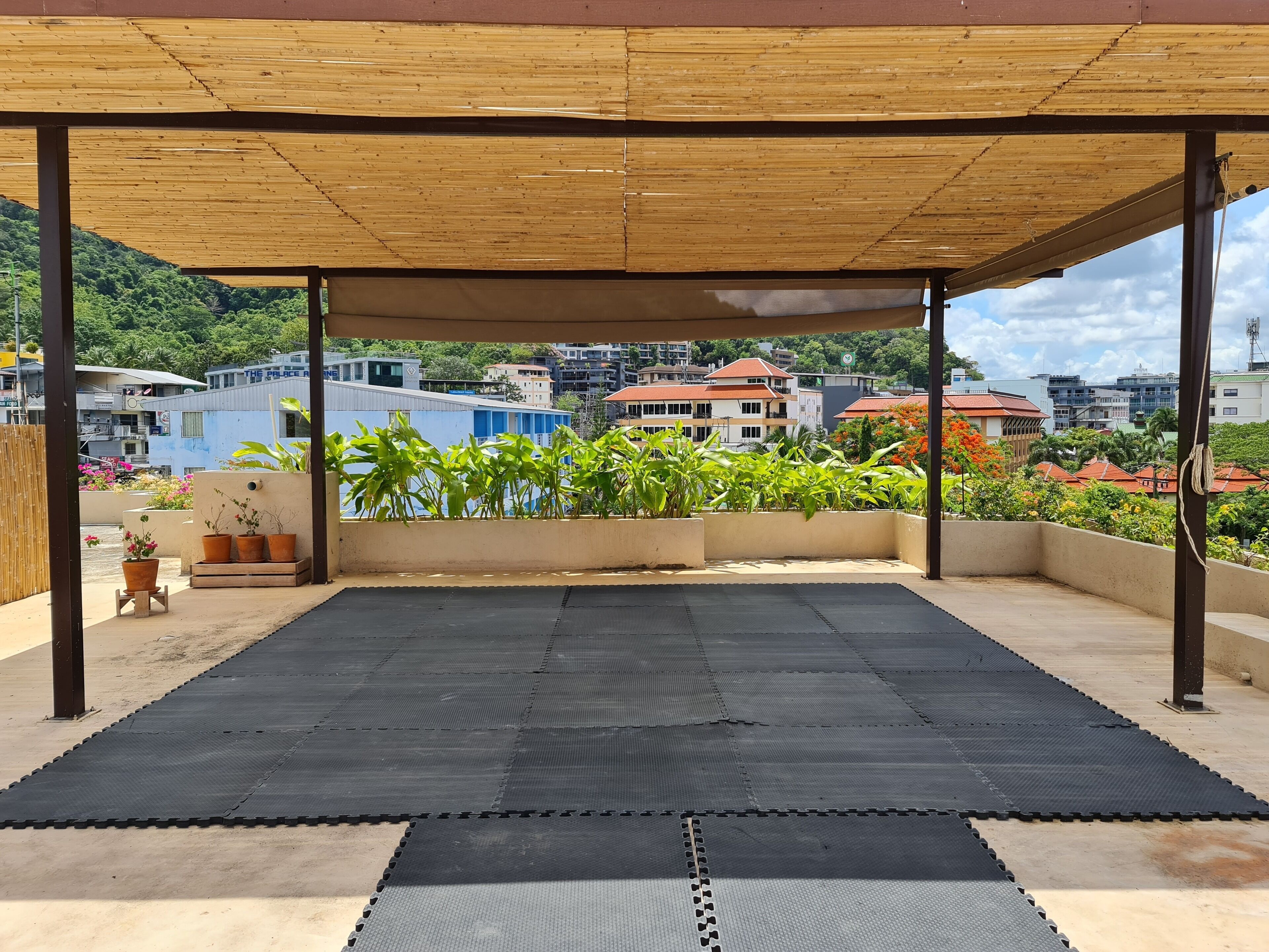 A covered outdoor area with a black rubber mat floor, surrounded by potted plants and a view of buildings in the background.