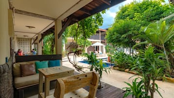 Un banco y una mesa de madera bajo una zona techada, con vista a una piscina y follaje exuberante.