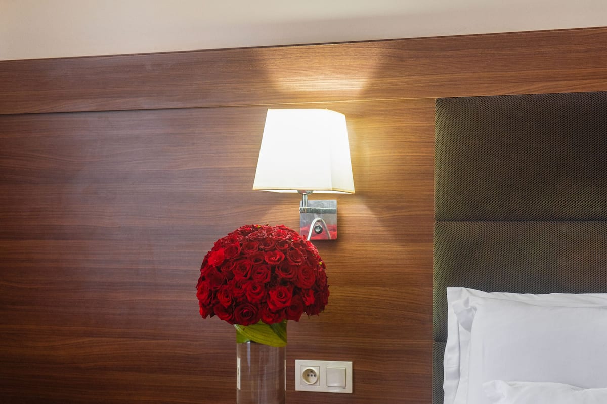A hotel room with a wooden headboard, a bedside table with a lamp, a vase of red roses, and a white bed with pillows.