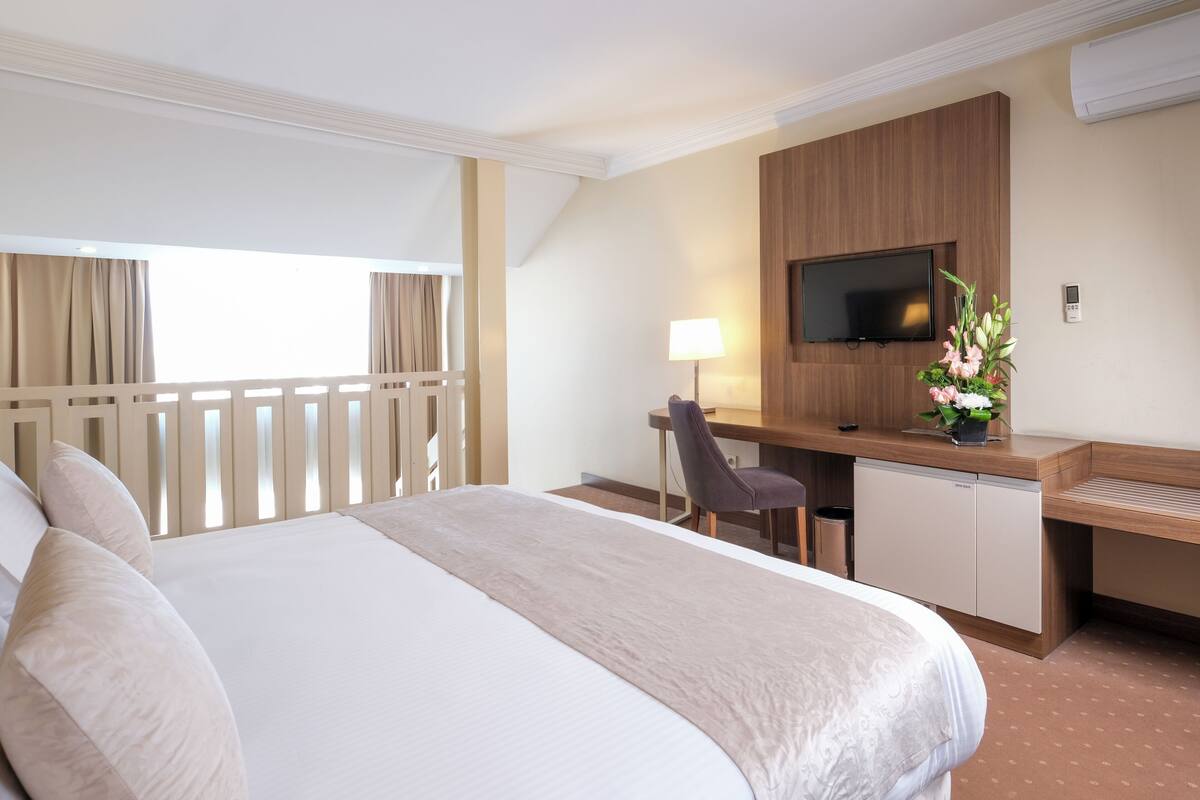 A hotel room with a large bed, a desk with a chair, a television, and a small desk with a lamp and flowers.