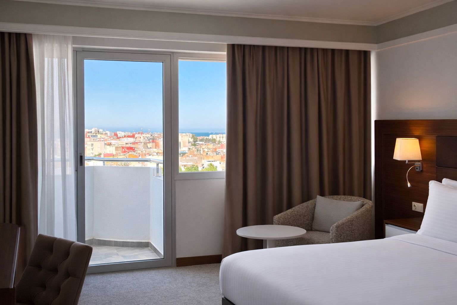 A hotel room with a bed, a chair, a small table, a nightstand, a lamp, and a view of the city through a large window.