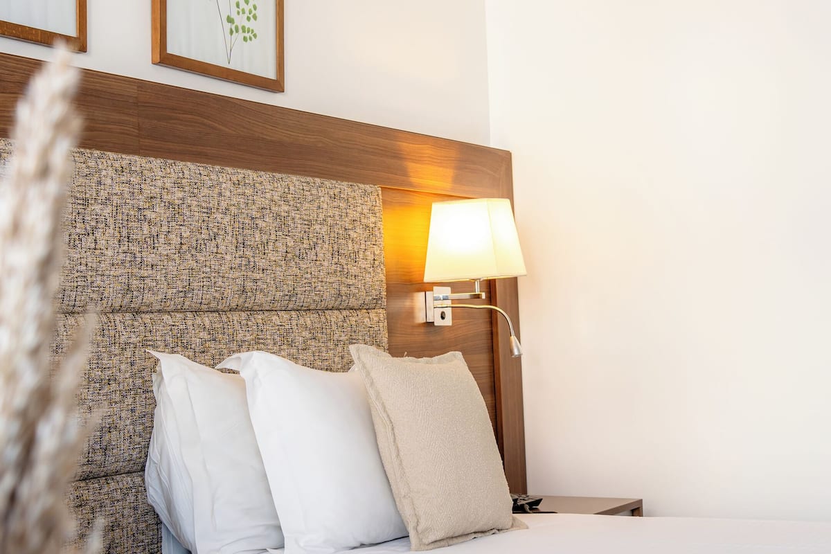 A hotel room with a large bed, a headboard with a textured pattern, a bedside lamp, and two framed botanical prints on the wall.