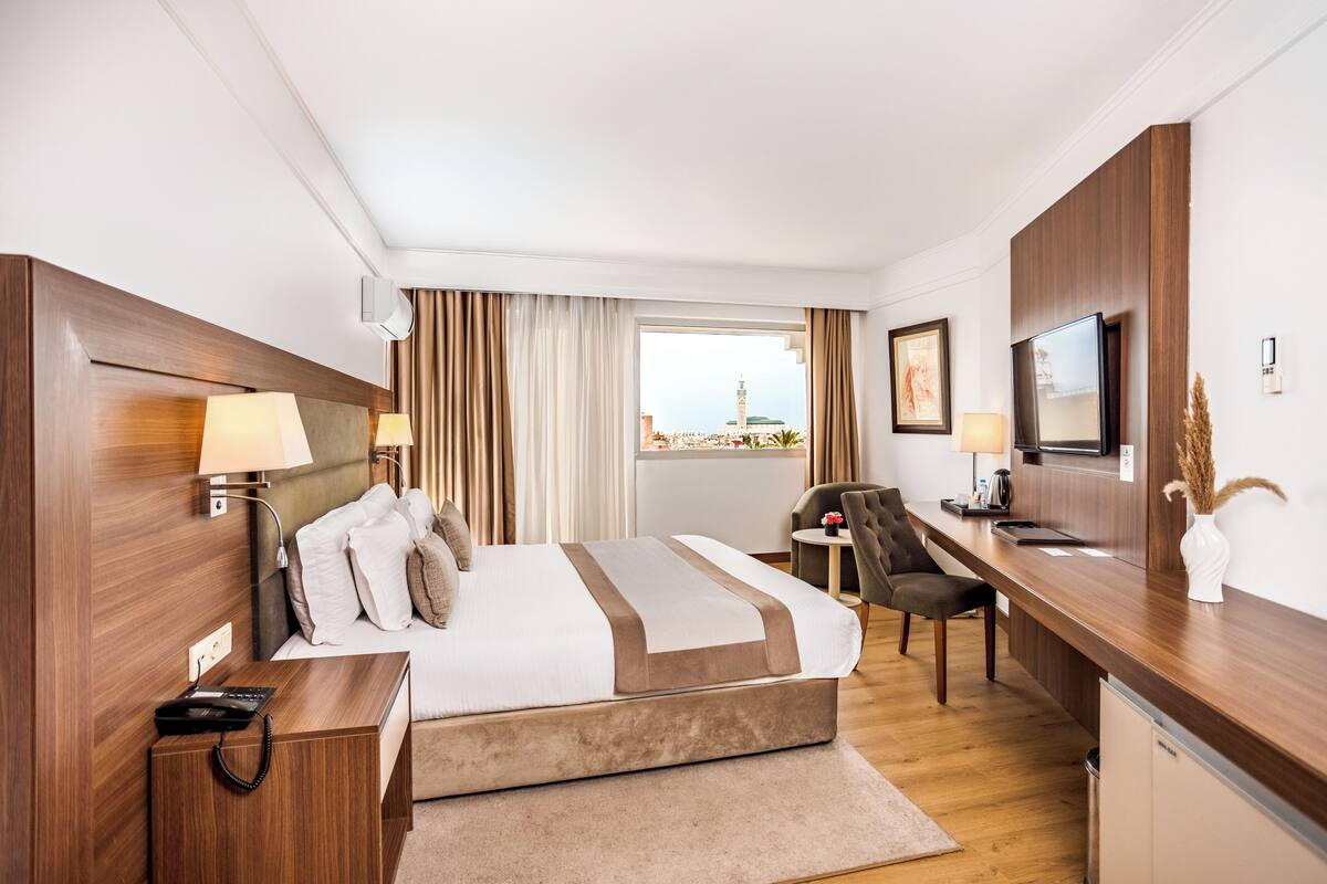 A modern hotel room with a large bed, a desk with a chair, a television, and a window with curtains.