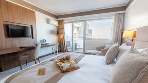 Habitación de hotel con cama, bandeja de desayuno, televisor, escritorio, silla y balcón con vistas.