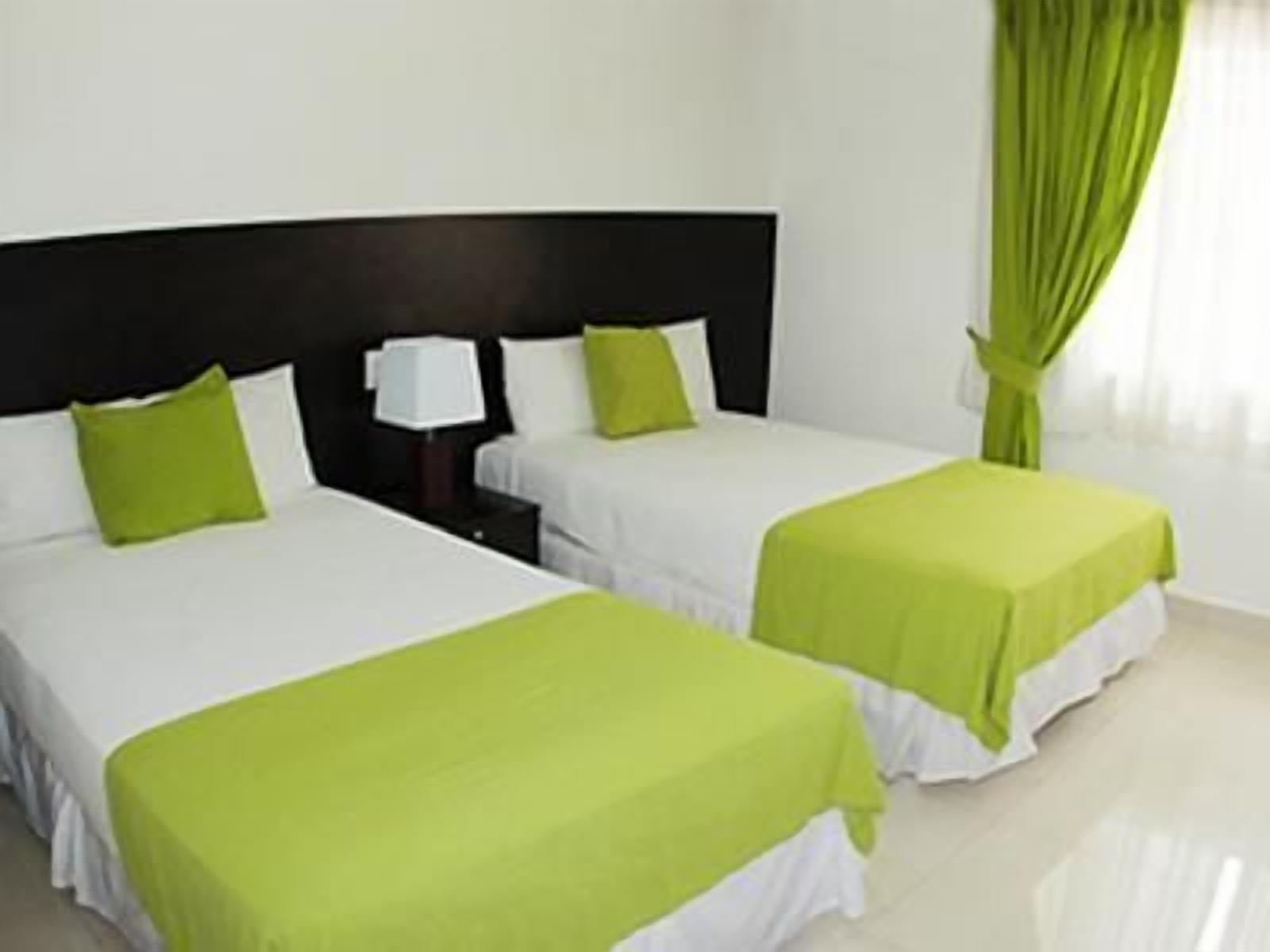 A hotel room with two beds, each with a white bedspread and green pillows, a nightstand with a lamp, and a window with green curtains.