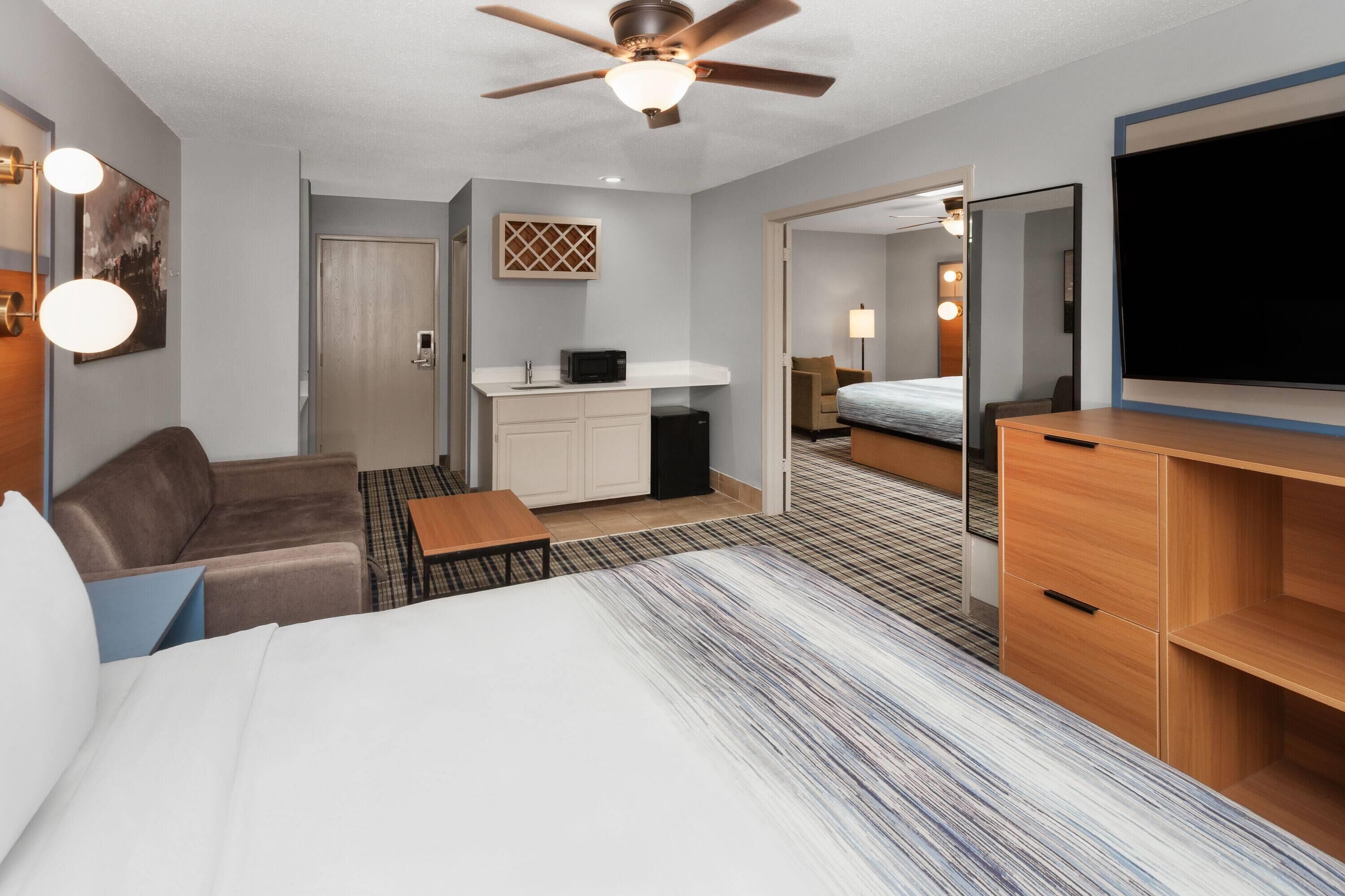 A hotel room with a bed, a ceiling fan, a desk, a chair, a sofa, a TV, a mirror, a door, and a small kitchenette.