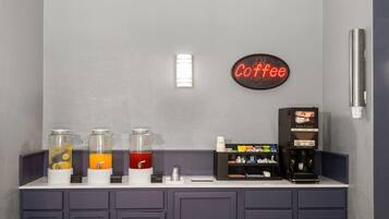 Una estación de café con dispensadores de agua, fruta y zumo de naranja, una máquina de café y un letrero de neón que dice 'Coffee'.