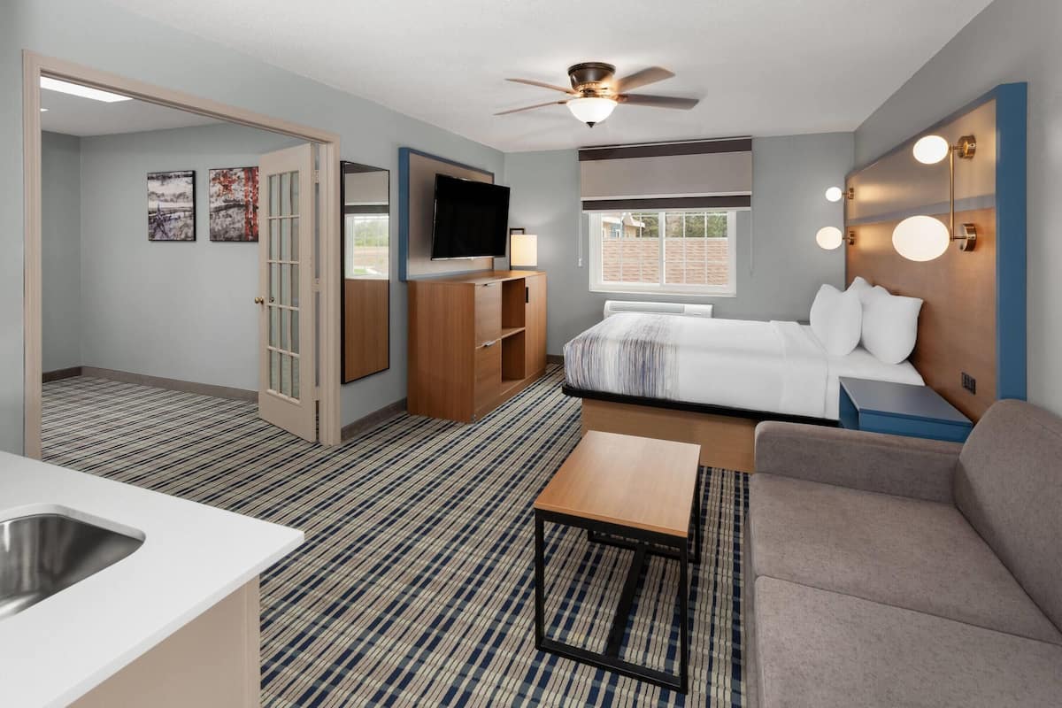 A hotel room with a bed, a sofa, a TV, a ceiling fan, a window, and a kitchenette.