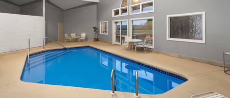 Una piscina interior con agua azul cristalina, rodeada de suelo color beige, y equipada con sillas metálicas junto a la piscina y una pequeña mesa.