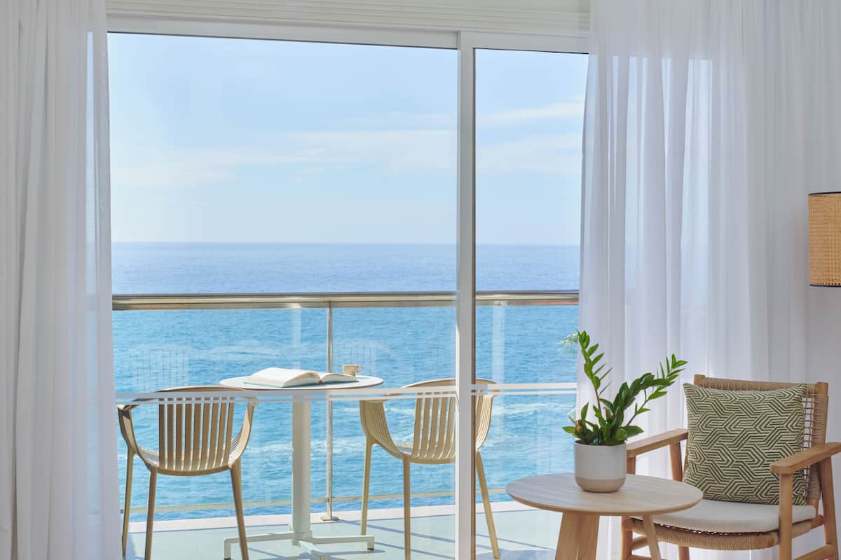 Un balcon avec vue sur la mer, deux chaises, une table avec un livre et une tasse, et une plante en pot.