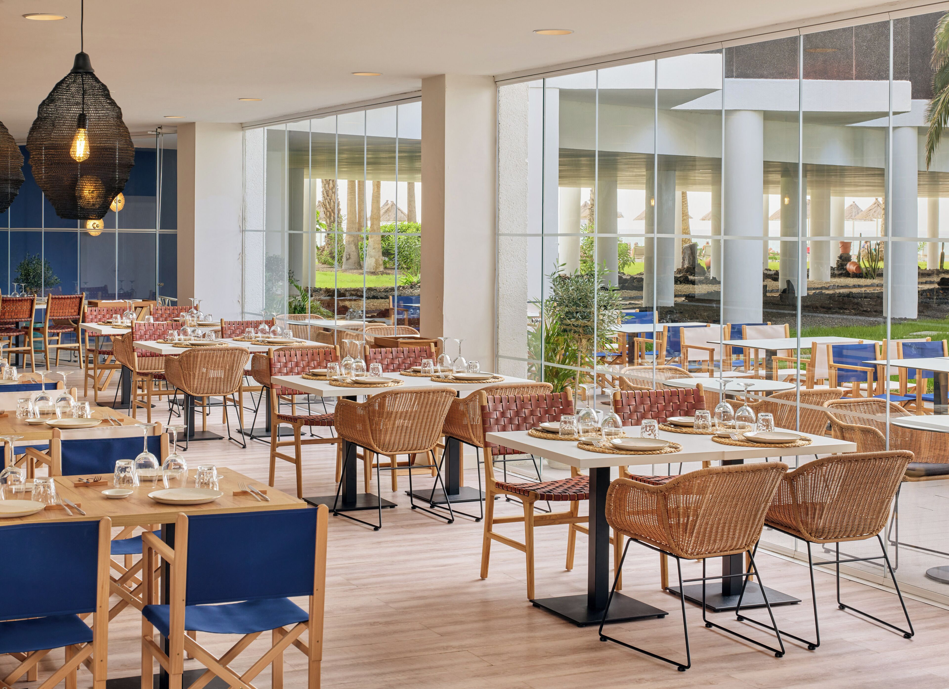 A spacious restaurant with high ceilings, large windows, and a mix of wooden and wicker furniture.
