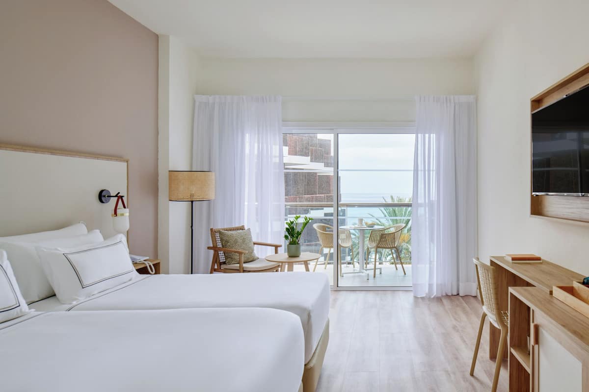 Une chambre d’hôtel avec deux lits, un bureau, une chaise, une télévision et un balcon donnant sur la mer.