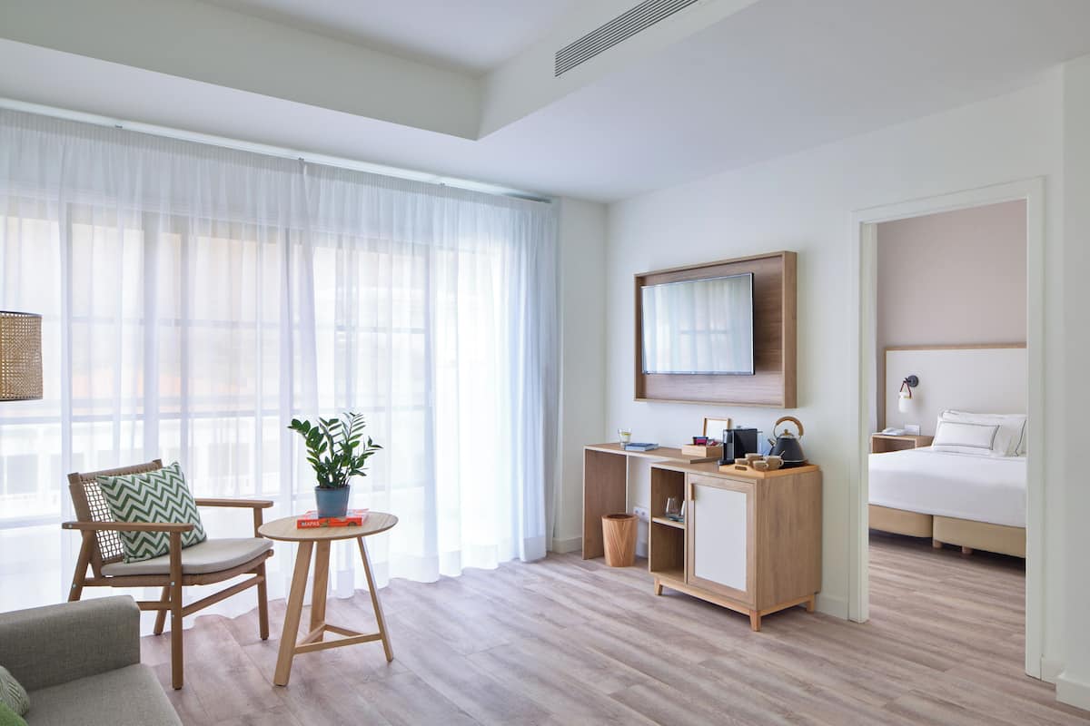 Une chambre d’hôtel moderne avec un bureau en bois, un lit, une chaise et une grande fenêtre avec des rideaux légers.