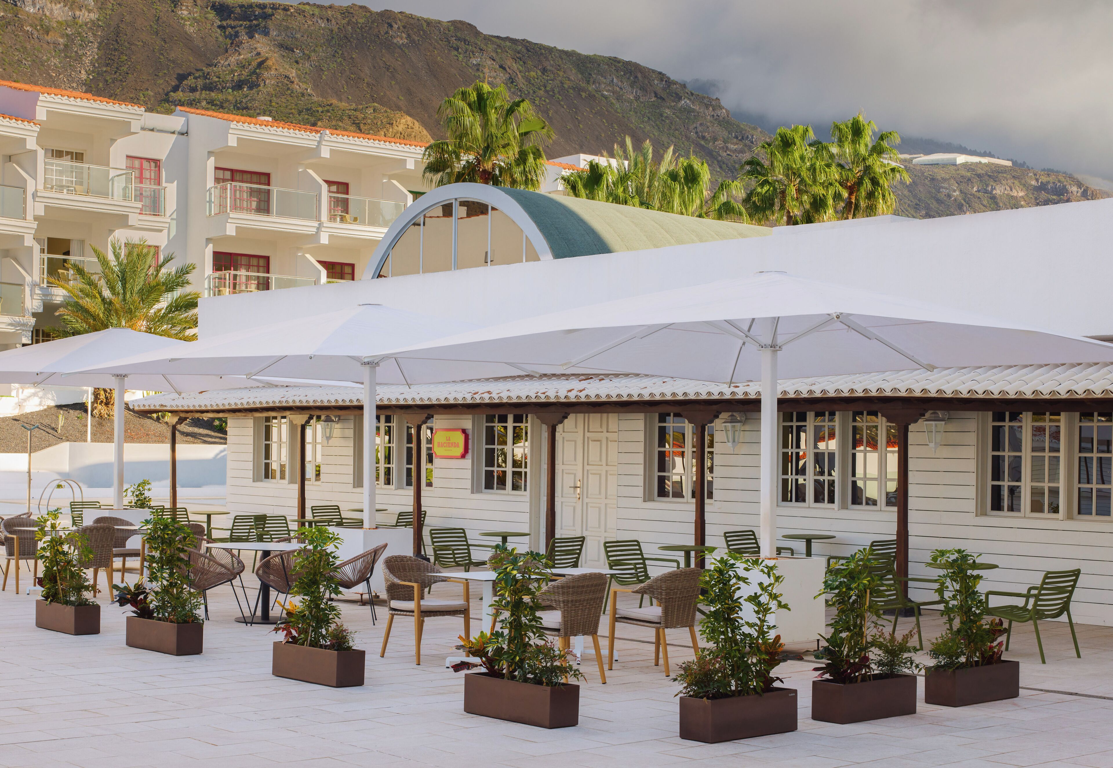 A seaside restaurant with outdoor seating, white umbrellas, and a view of the mountains.