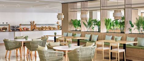 A modern dining area with wicker chairs and tables, a kitchen in the background, and a large window with plants.