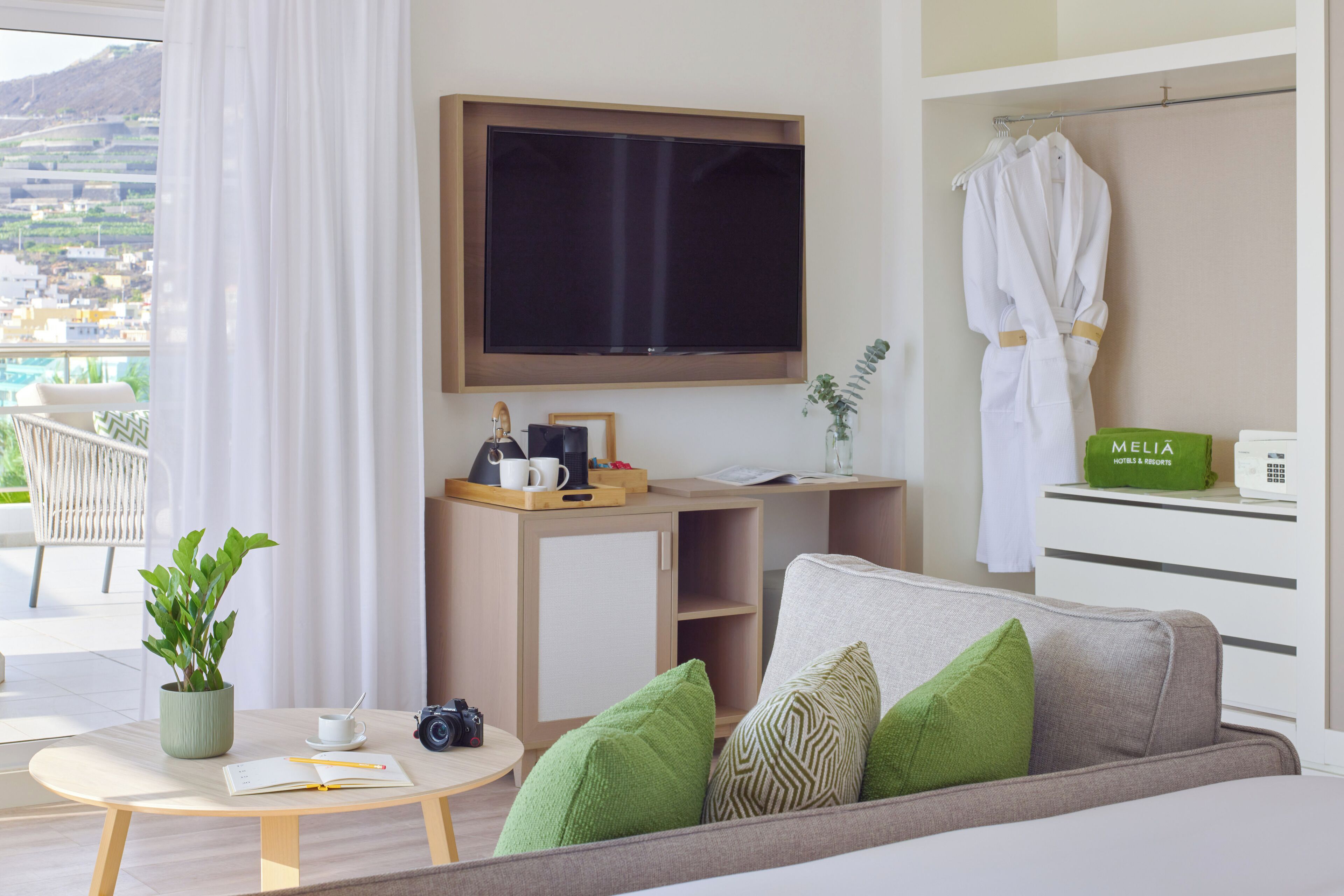 Une chambre d’hôtel moderne équipée d’un grand téléviseur à écran plat, d’un canapé avec des coussins verts, d’une petite table ronde agrémentée d’une plante, d’une machine à café et d’un appareil photo.