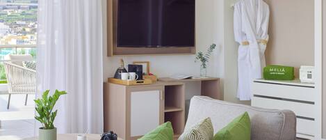 A modern hotel room with a large flat-screen TV, a sofa with green cushions, a small round table with a plant, a coffee maker, and a camera.