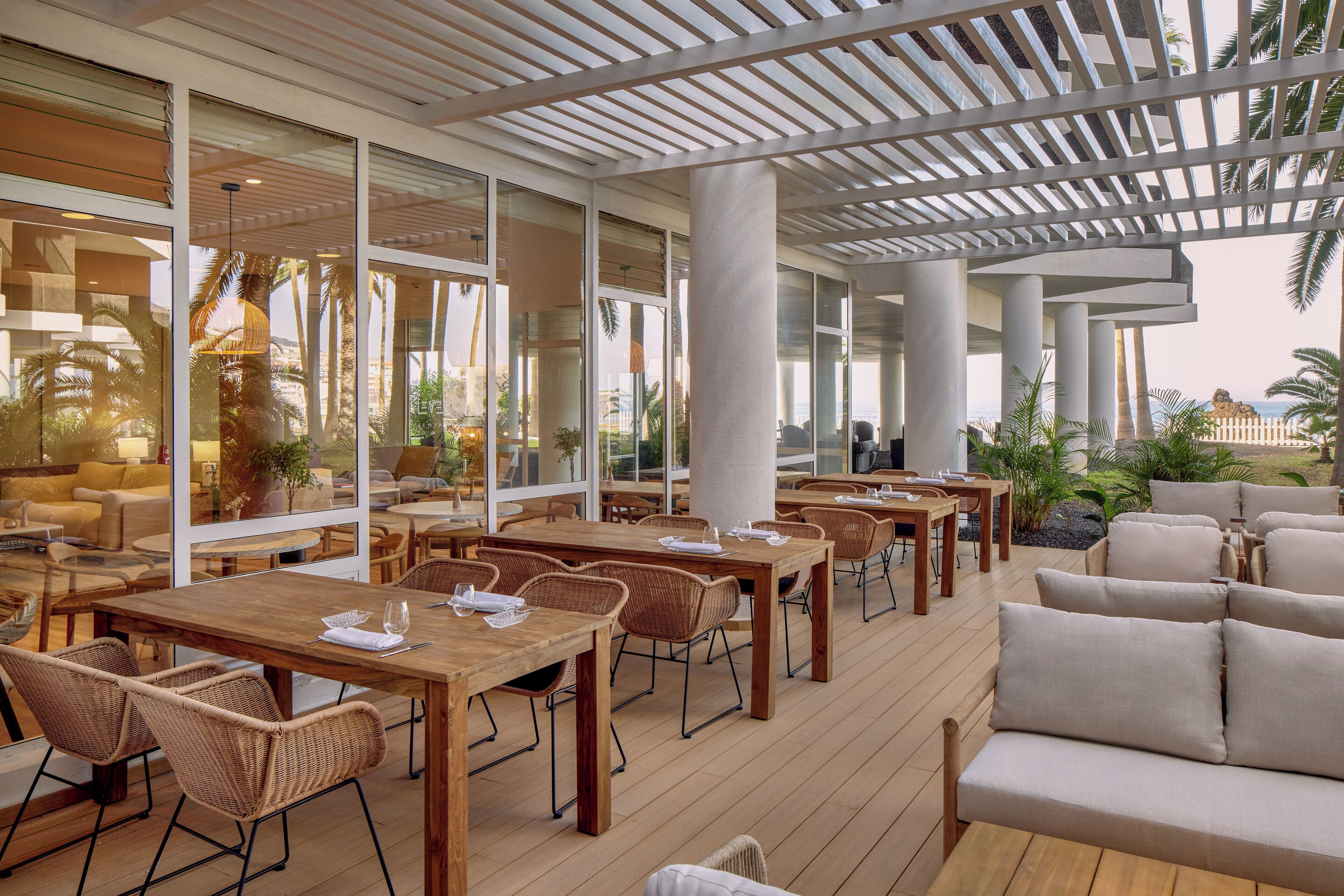 Un área de comedor al aire libre con mesas de madera y sillas de mimbre, ventanas amplias y vistas a palmeras.