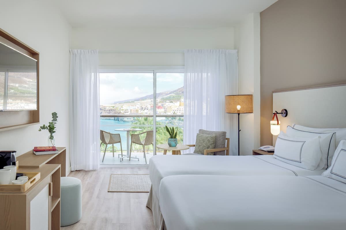Une chambre d’hôtel avec un grand lit, un bureau avec une chaise, une petite table avec des chaises, une lampe et une vue sur la ville et les collines par la fenêtre.