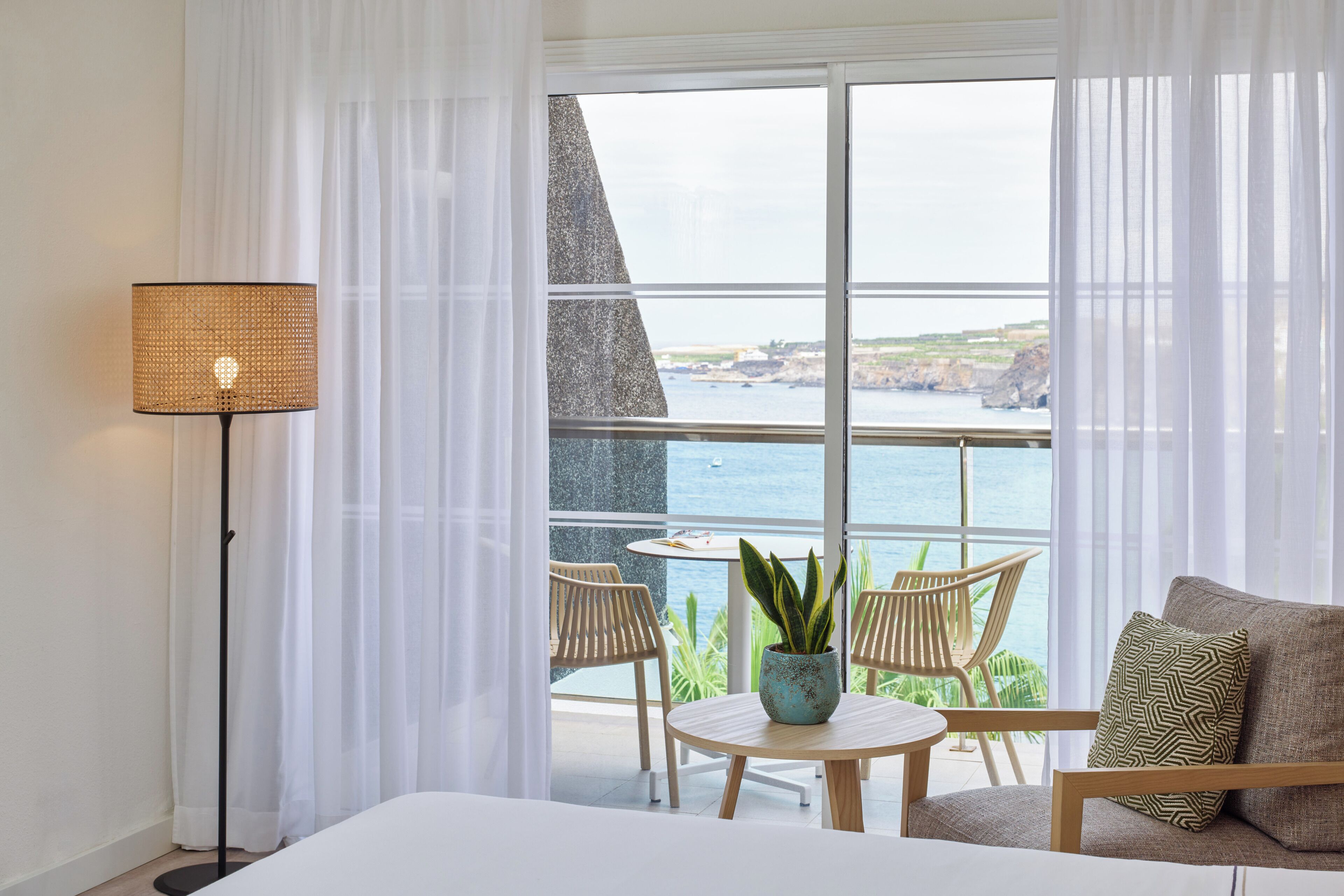 A hotel room with a balcony, a bed, a table with chairs, a potted plant, and a lamp.