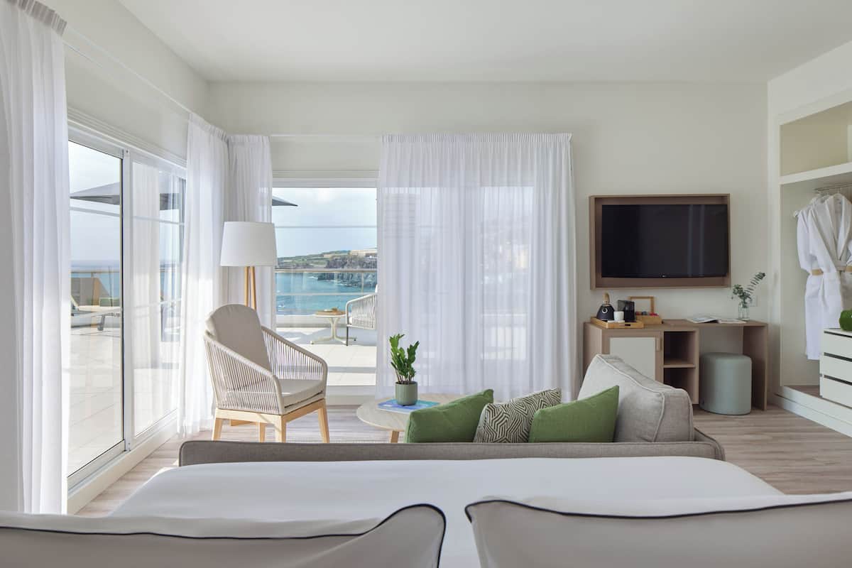 Une chambre d’hôtel moderne dotée d’un balcon, d’une télévision, d’un canapé avec des coussins, d’un fauteuil, d’une petite table avec une lampe et d’un bureau agrémenté d’une plante.