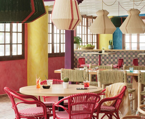 Un comedor con mesas redondas, sillas de mimbre rojo y lámparas colgantes.