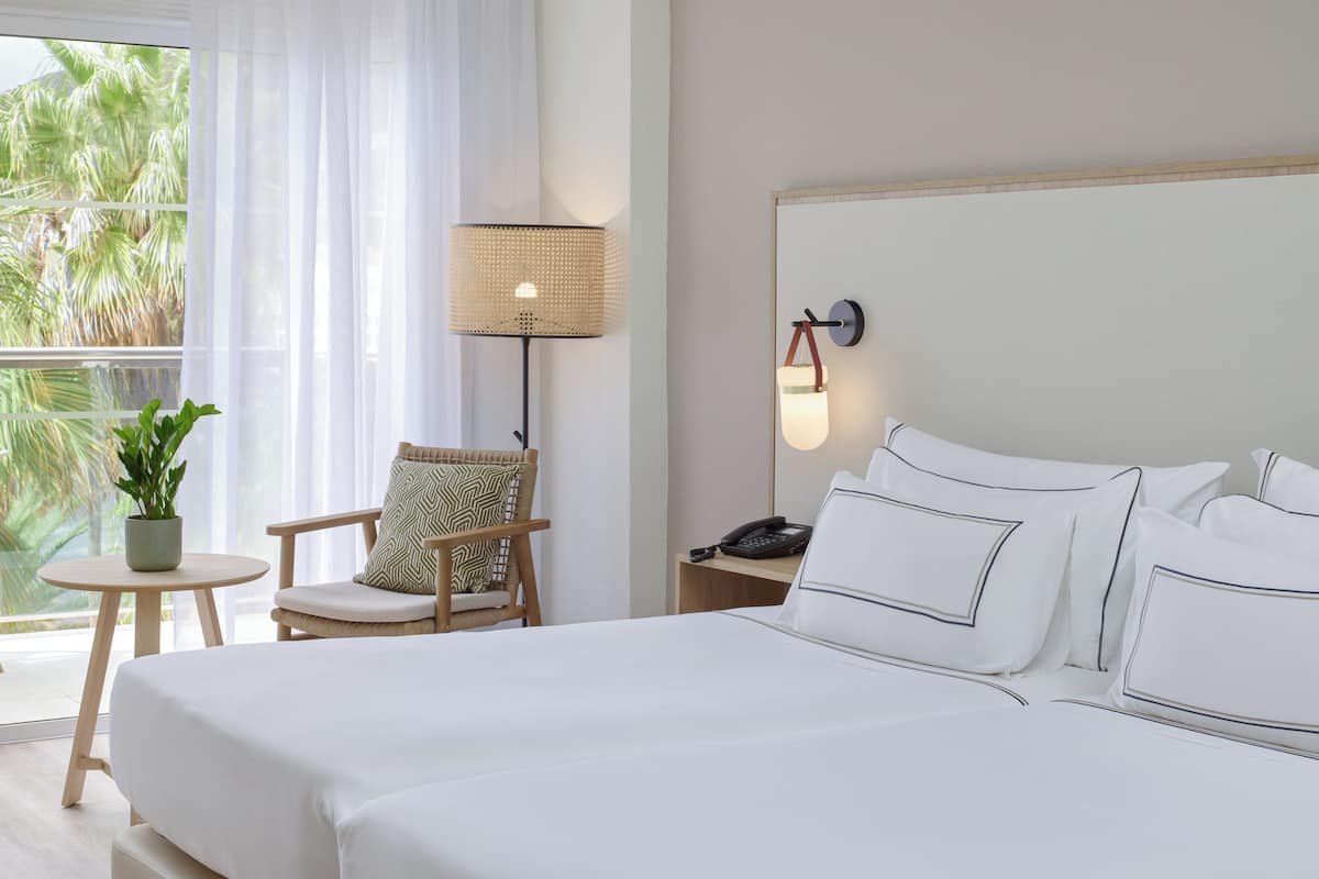 Une chambre d’hôtel avec un grand lit, une chaise, une petite table avec une plante, une lampe et une table de chevet avec un téléphone.