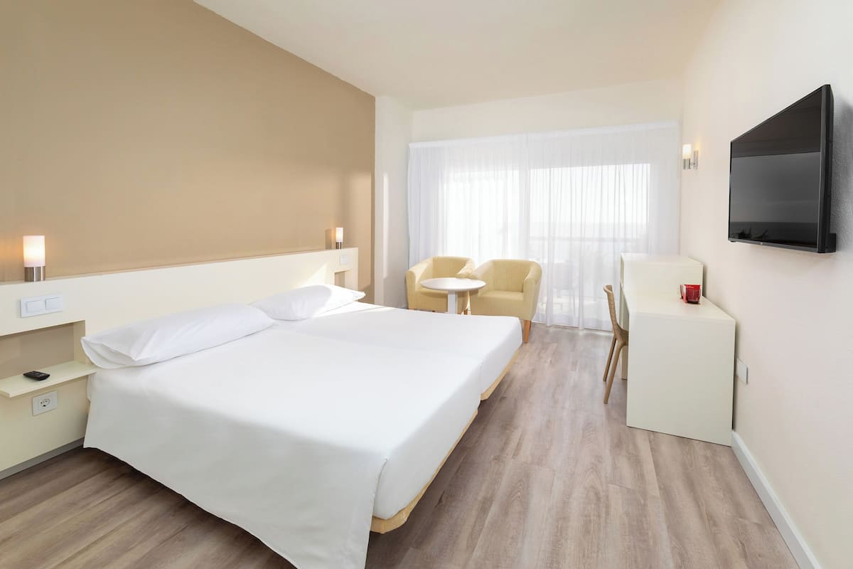 Une chambre d’hôtel avec un grand lit, une petite table, une chaise, un téléviseur et une fenêtre avec des rideaux légers.