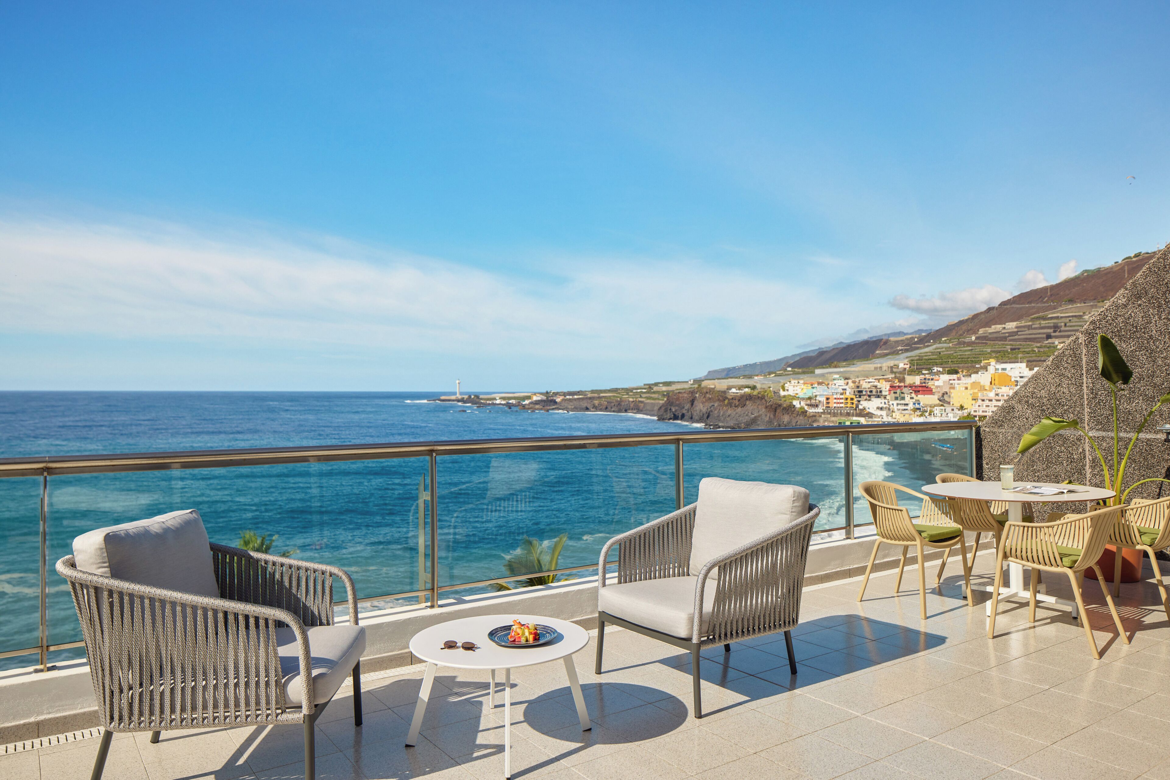 A balcony with wicker chairs and a table, overlooking a coastal view with buildings in the distance.
