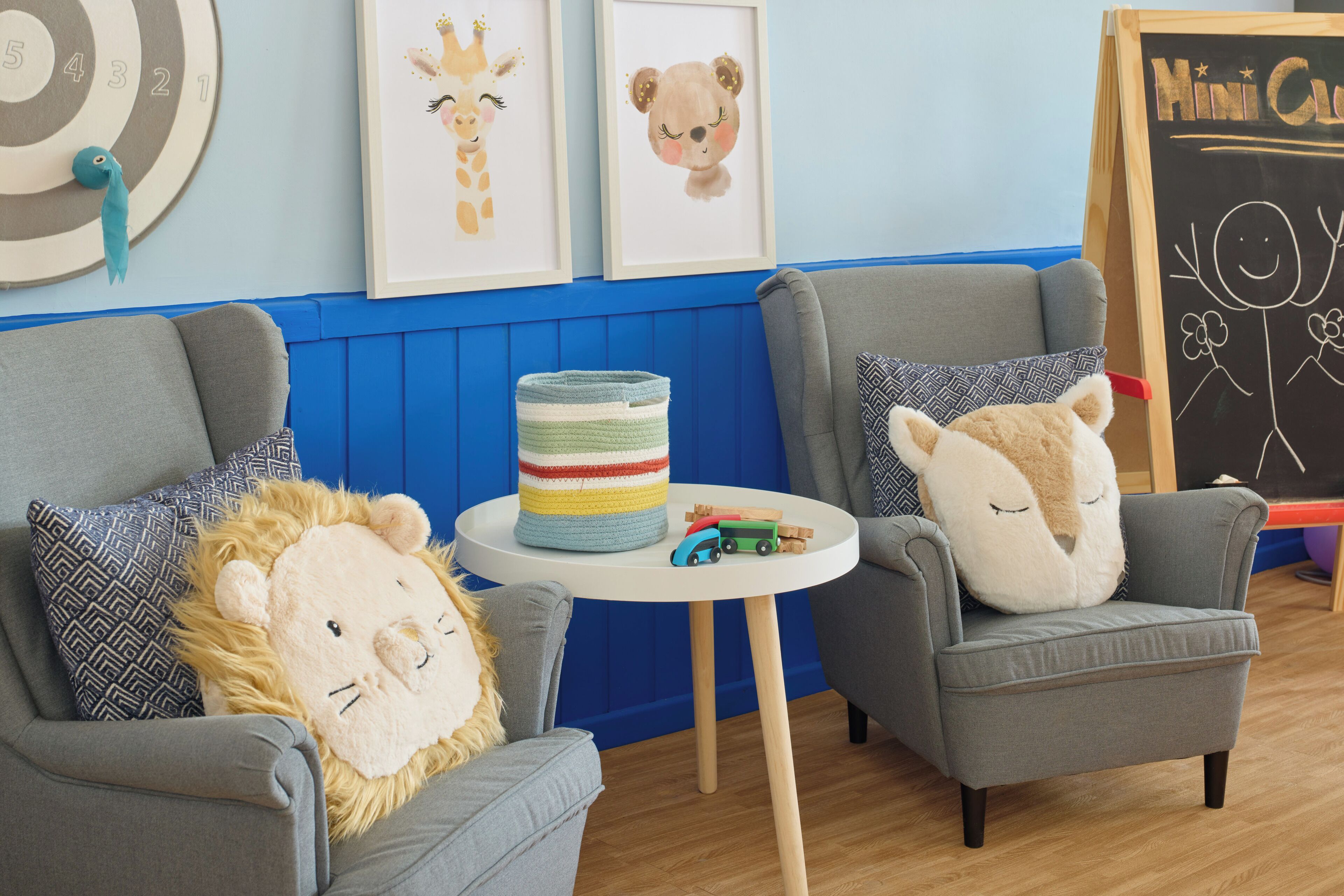 Ein Kinderspielzimmer mit zwei Sesseln, einem kleinen Tisch mit Spielzeug sowie Wanddekorationen, darunter gerahmte Tierbilder und eine Tafel mit Zeichnungen.