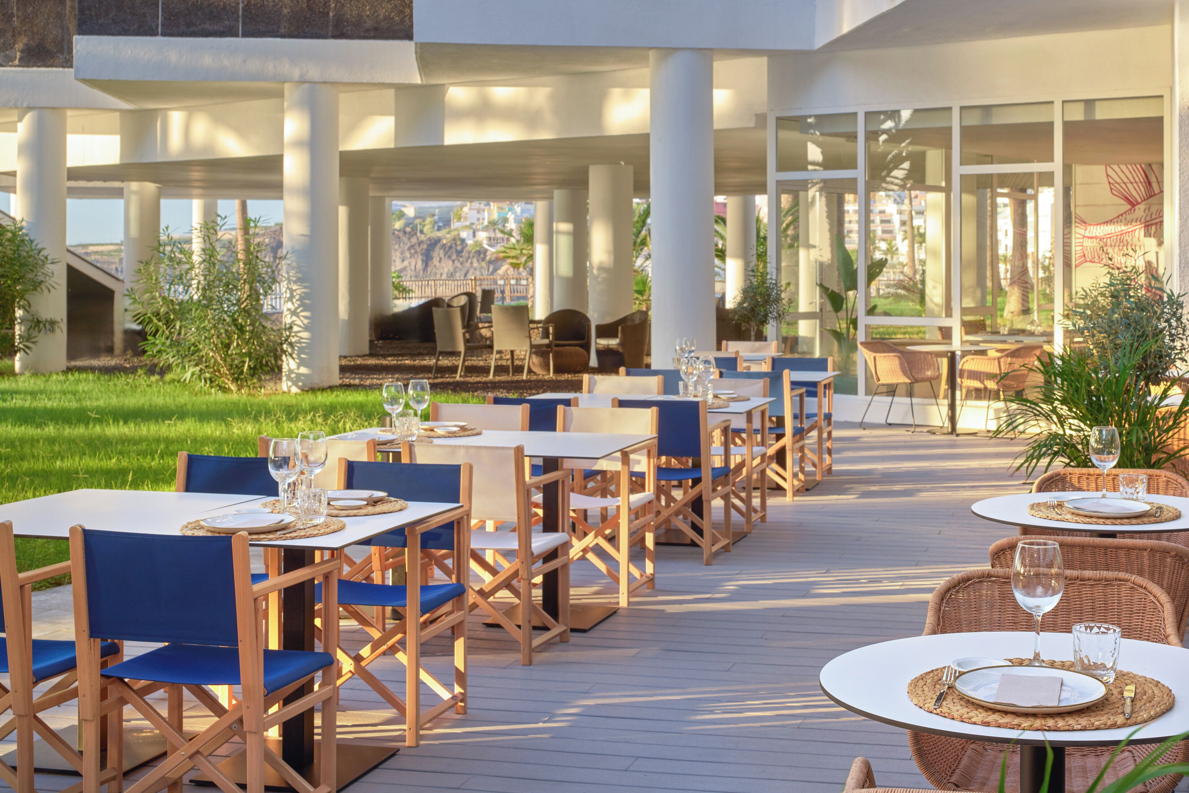 An outdoor dining area with tables and chairs, surrounded by greenery and a view of a hillside.