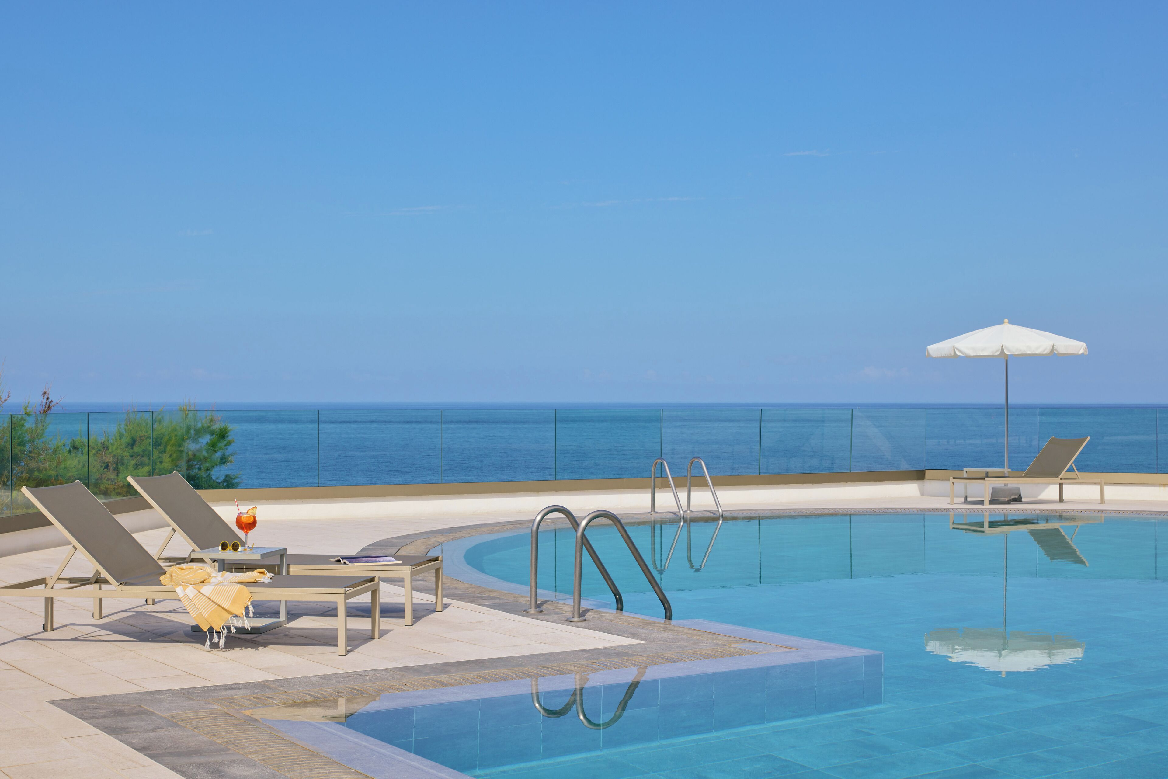 Ein Pool mit Liegestühlen und Sonnenschirmen, mit Blick auf das klare Meer und einer Metallleiter.