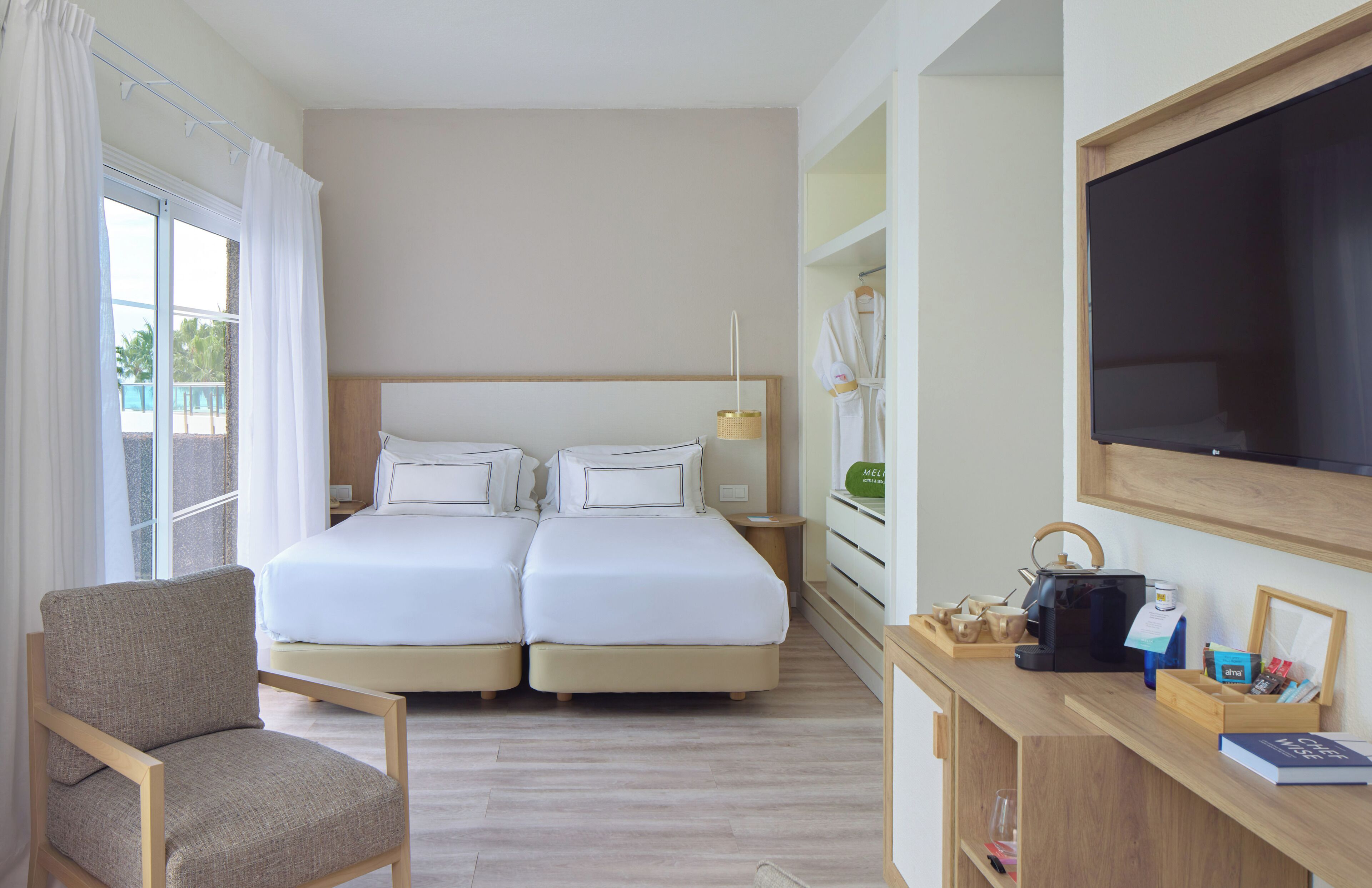 A hotel room with a double bed, a chair, a desk with a flat-screen TV, and a window with curtains.