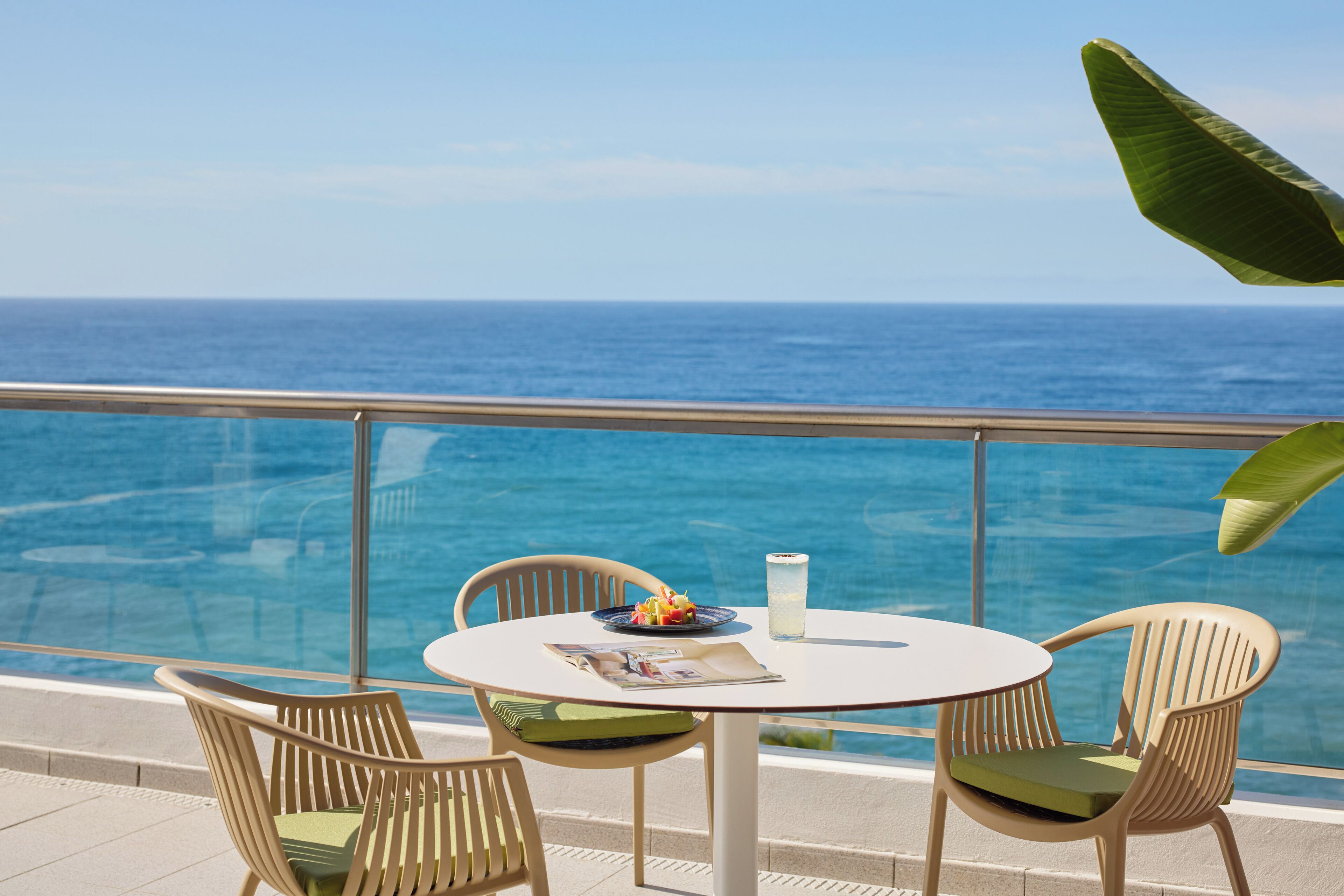 A round table with two chairs and a glass railing overlooking a pool and the ocean.