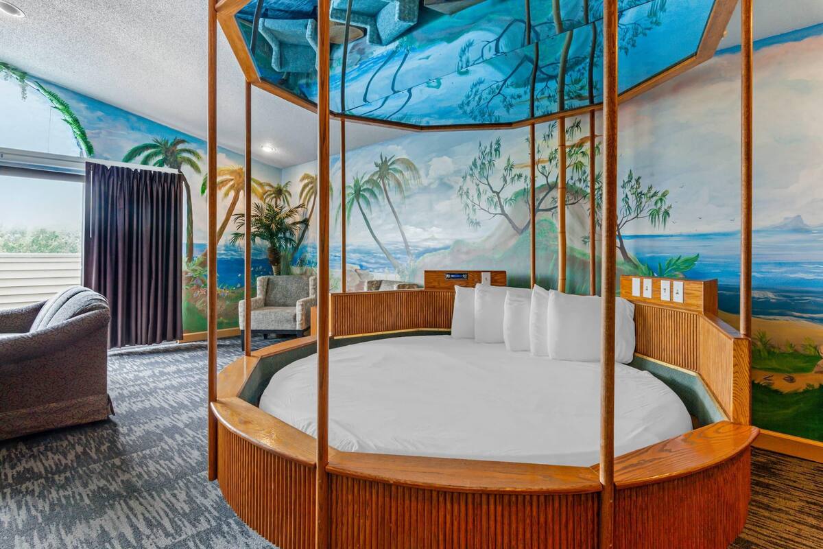A circular bed with a wooden frame, a view of the ocean through a window, and a tropical mural on the wall.