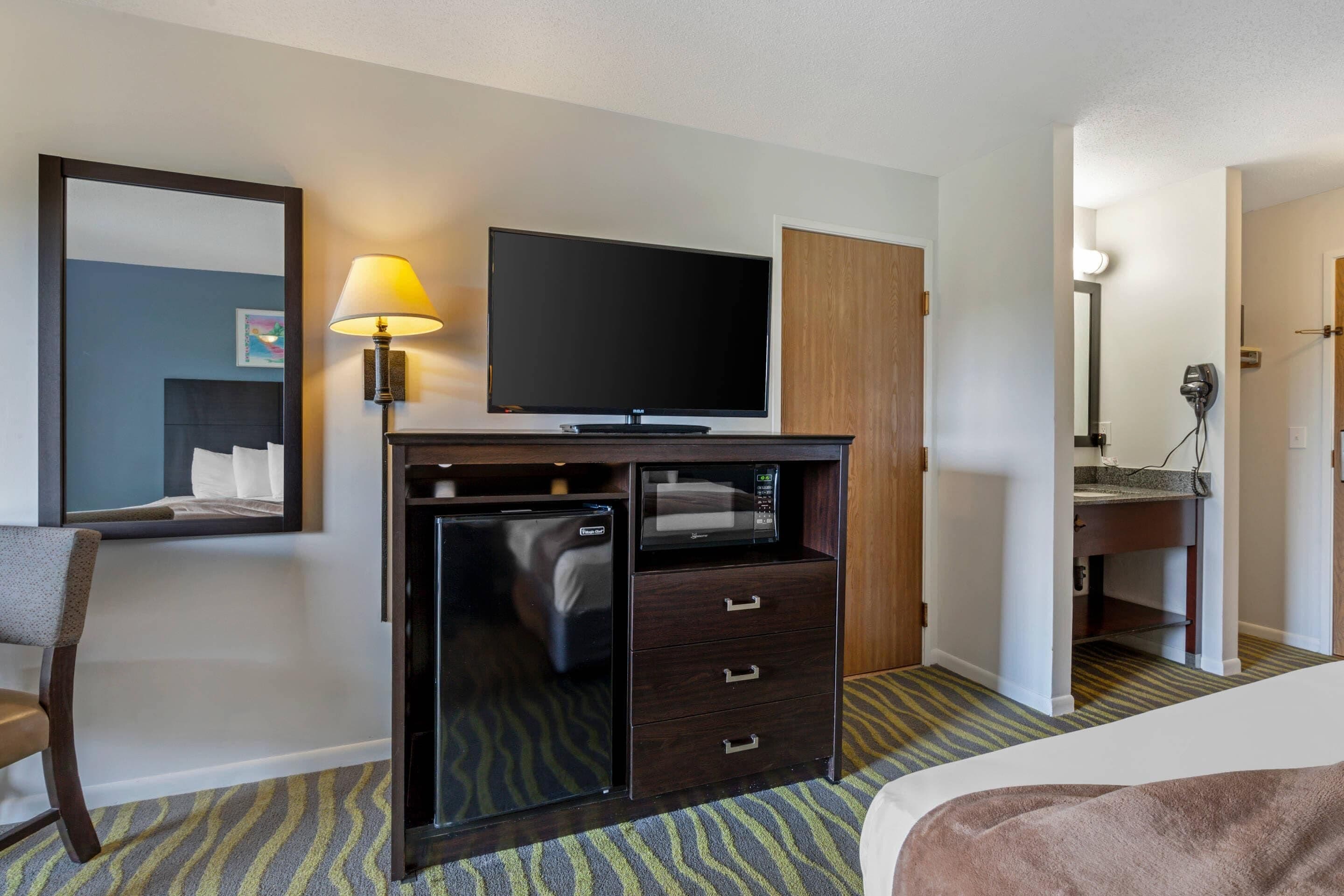 A hotel room with a bed, a desk with a television, a microwave, a hairdryer, and a mirror.