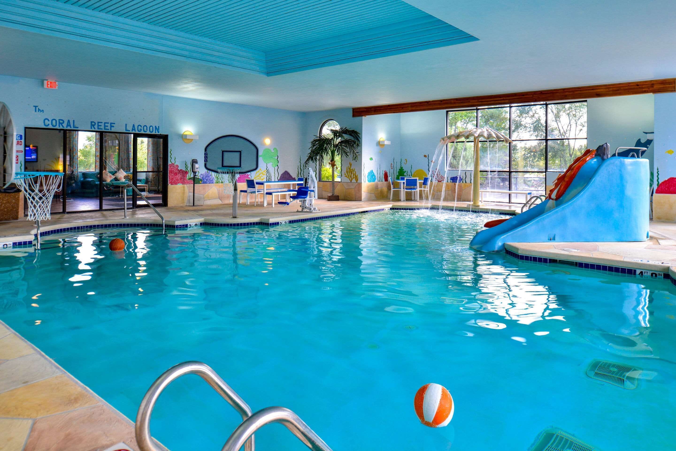 An indoor swimming pool with a basketball hoop, a slide, and a seating area with tables and chairs.