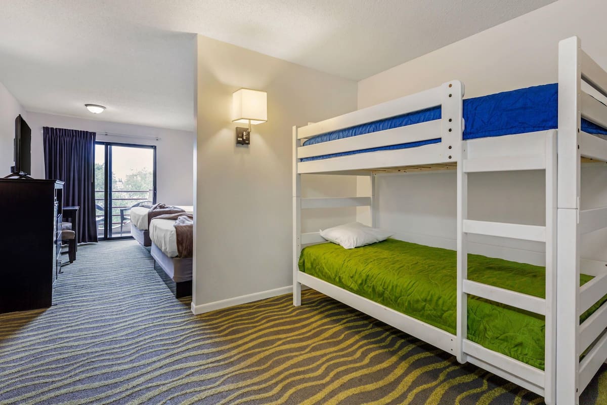 A hotel room with a bunk bed, a desk with a chair, a lamp, and a view of the outdoors through a large window.
