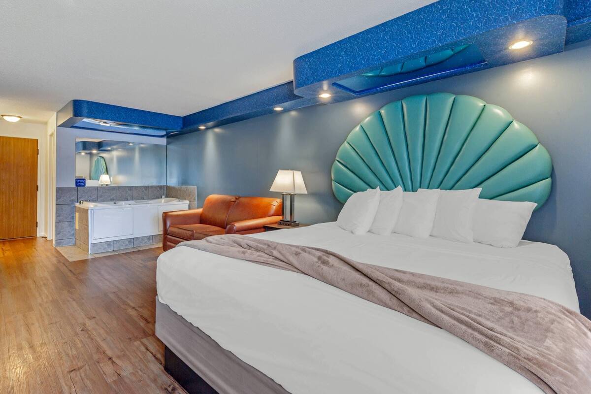 A modern hotel room with a large bed, a bathroom with a bathtub, and a seating area with a chair.