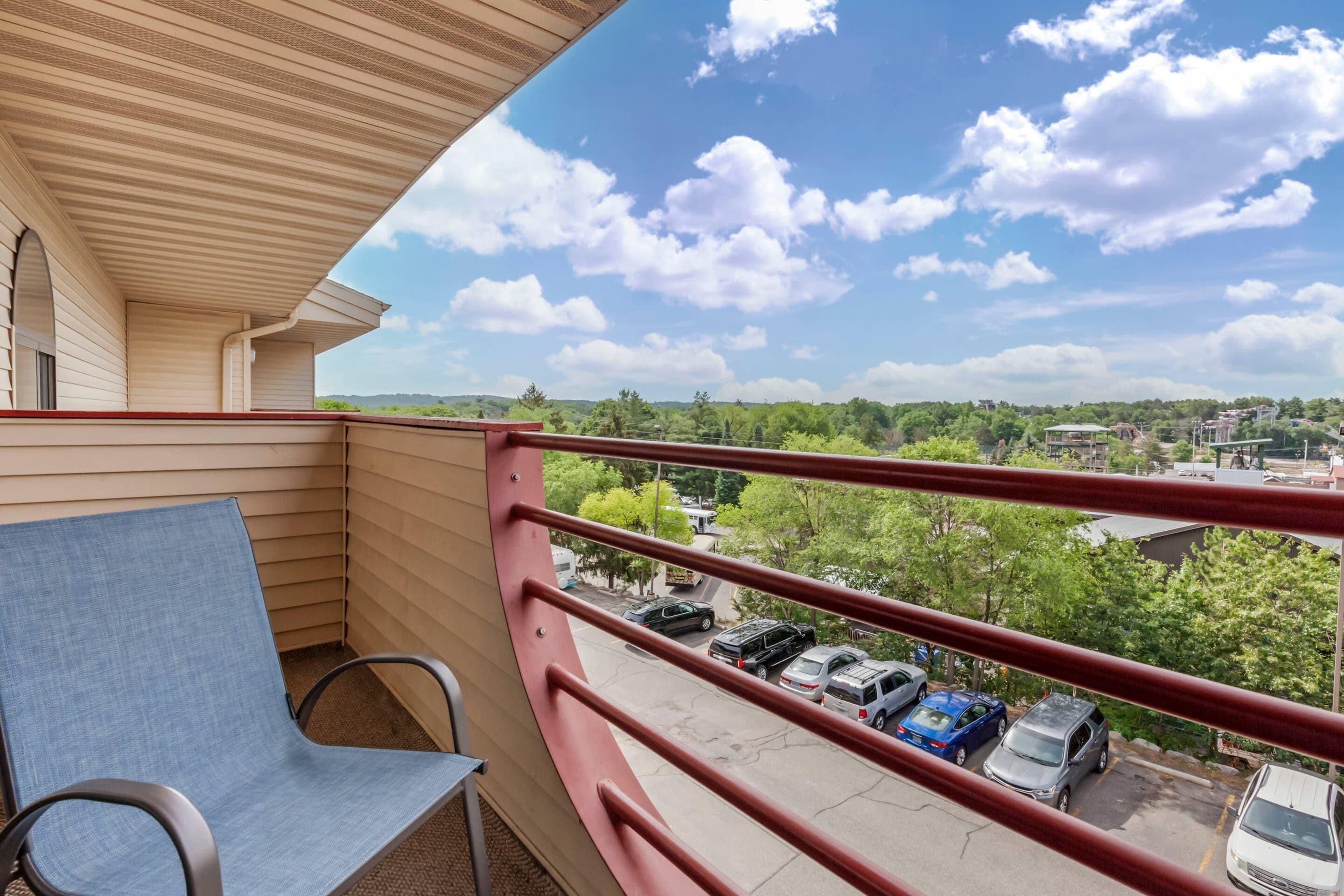 A balcony with a red railing, a blue chair, and a view of parked cars and greenery.
