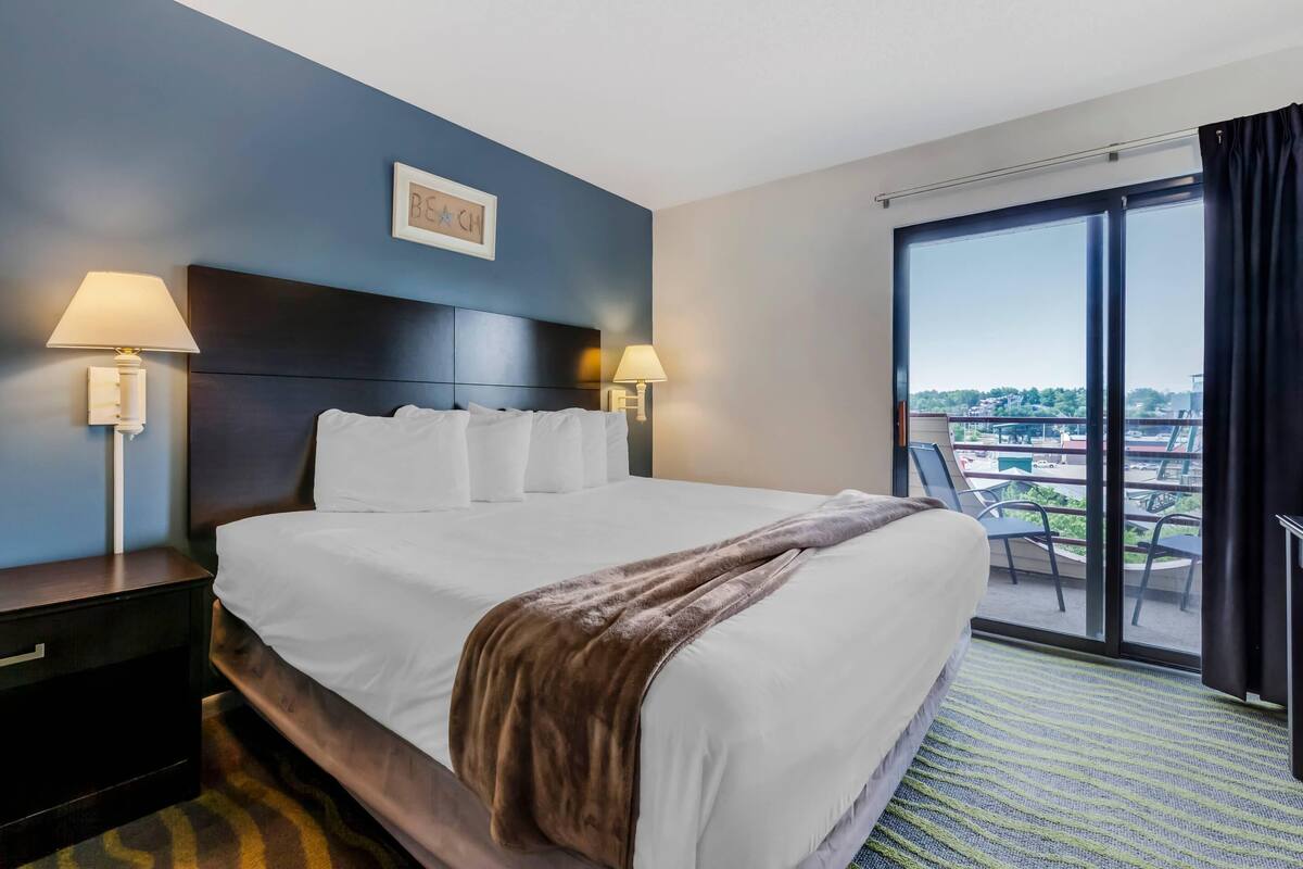 A hotel room with a large bed, two bedside lamps, a dark headboard, and a view of the outdoors through a sliding glass door.