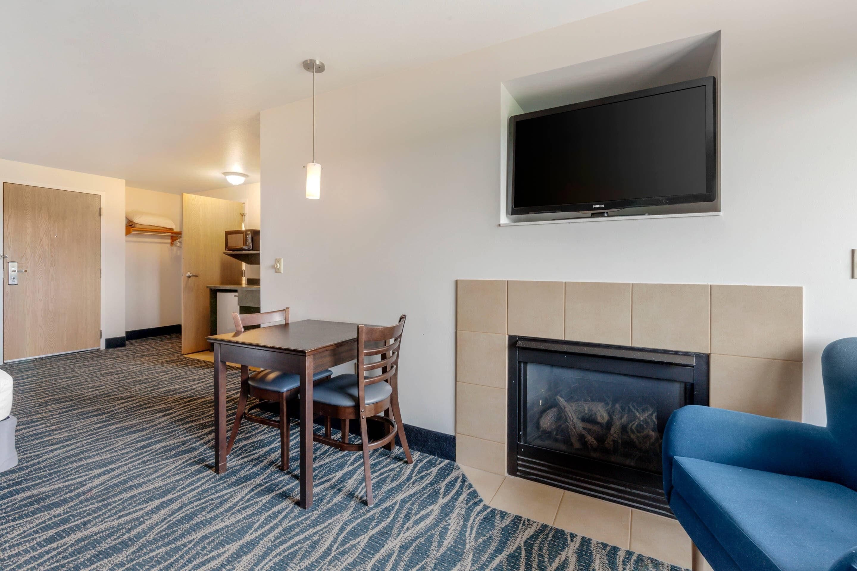 Habitación de hotel con televisión montada en la pared, chimenea, zona de comedor con mesa y sillas, y suelo alfombrado.