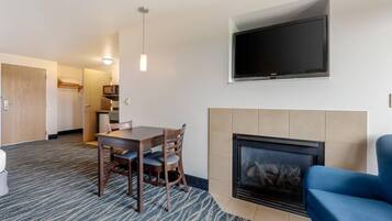 Habitación de hotel con televisión montada en la pared, chimenea, zona de comedor con mesa y sillas, y suelo alfombrado.