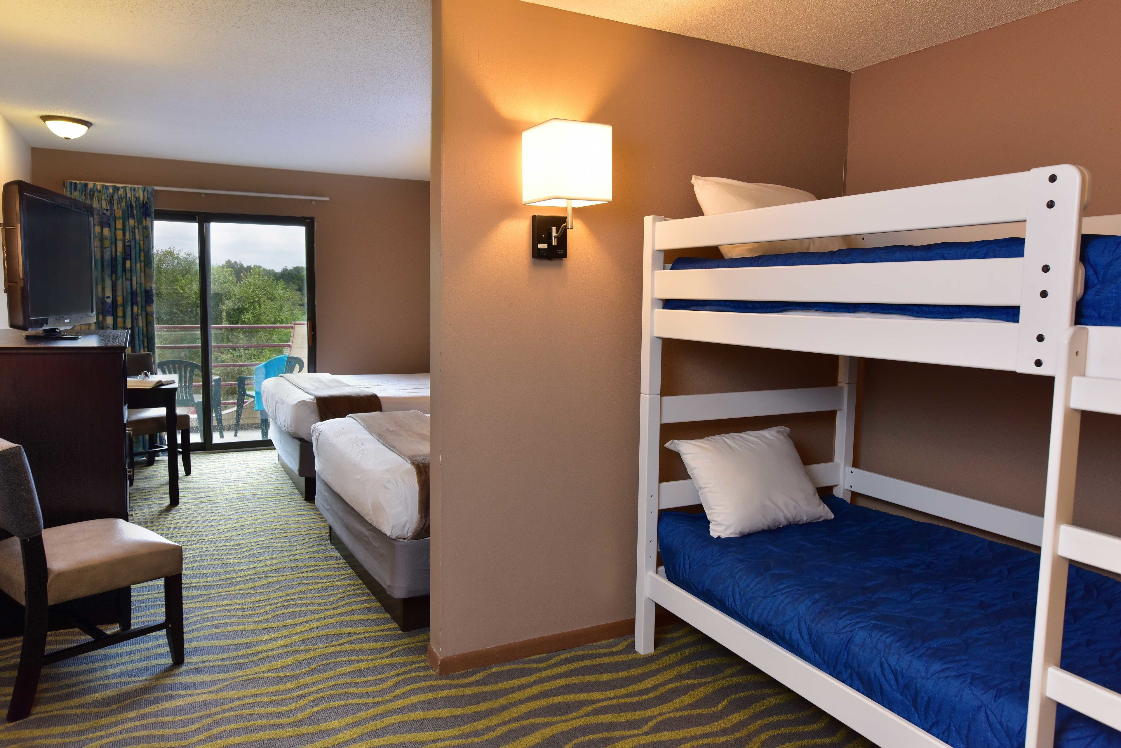 A hotel room with a bunk bed, a desk with a chair, a television, and a balcony with a view of trees.