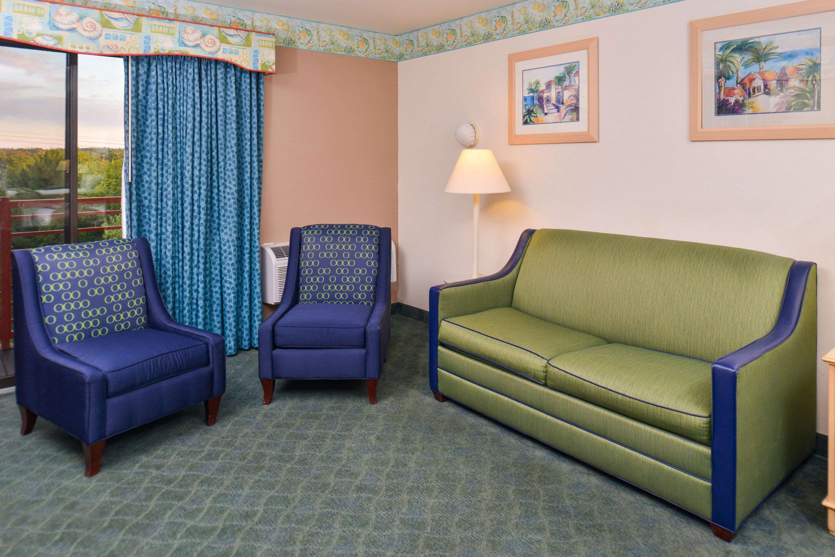 Une pièce avec un canapé vert, deux fauteuils à motifs, une fenêtre avec des rideaux bleus et deux tableaux encadrés accrochés au mur.