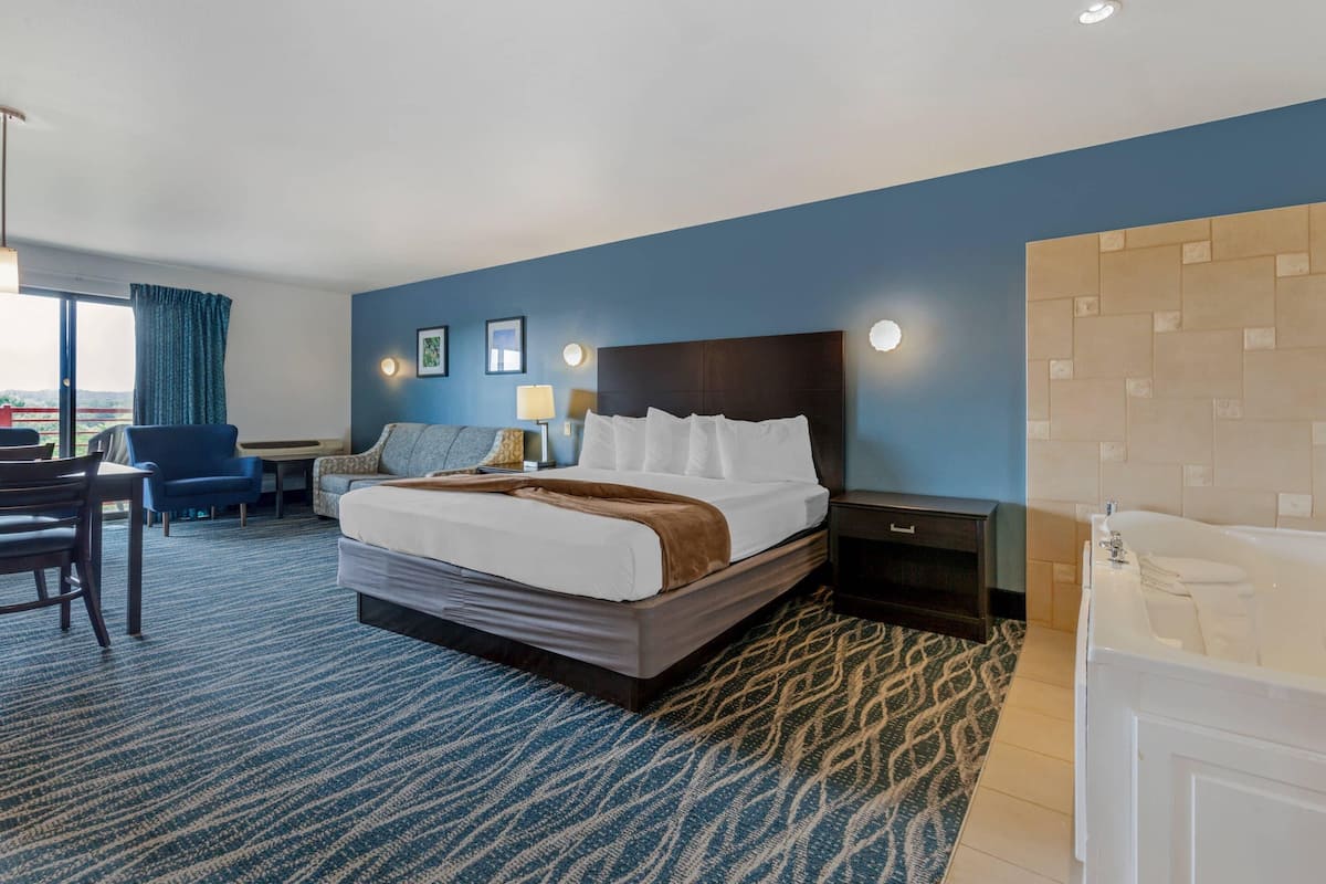 A hotel room with a large bed, a desk, a chair, a desk lamp, a nightstand, and a bathroom with a bathtub.