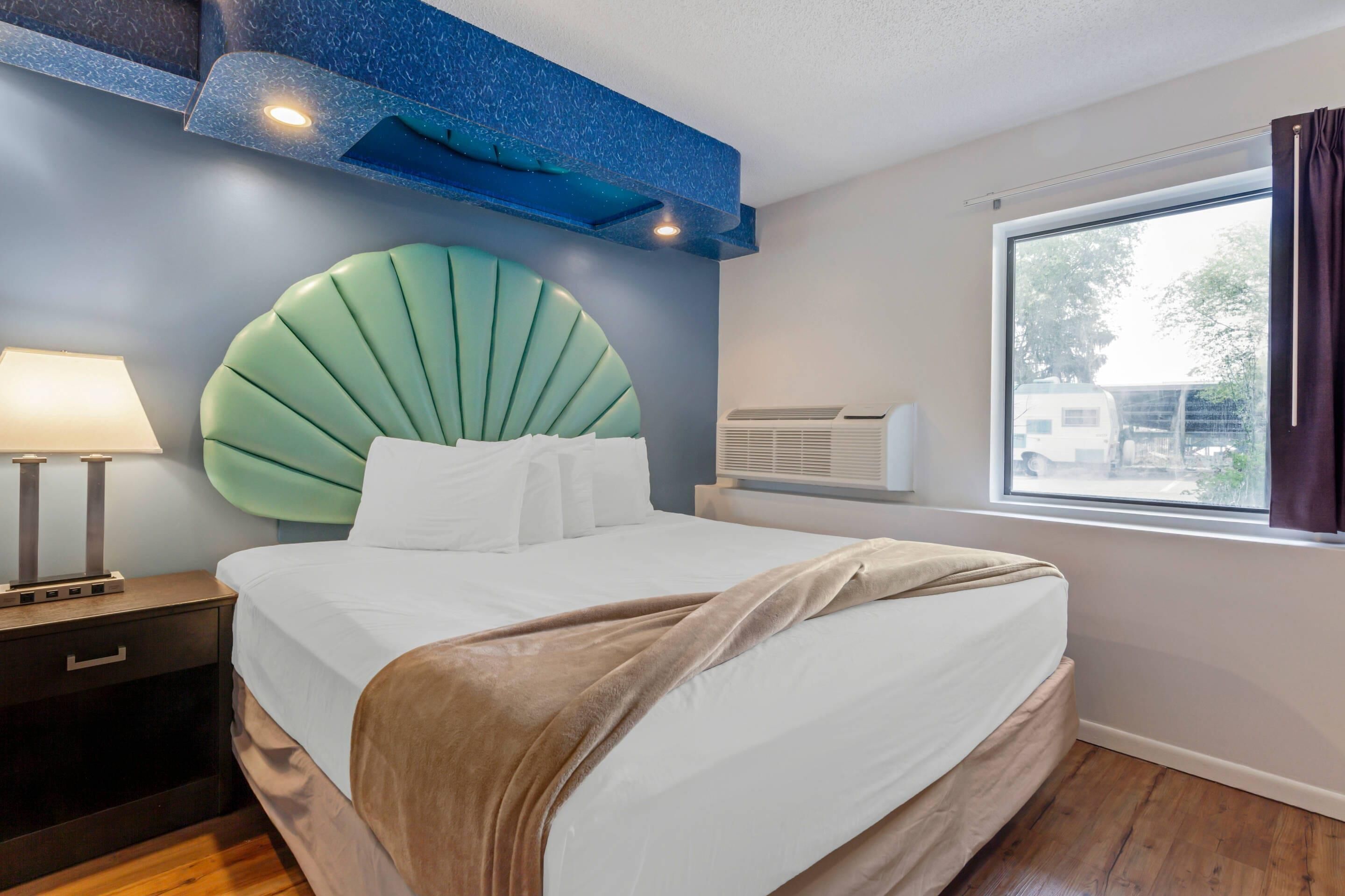 A modern hotel room with a large bed, a headboard with a green fan-like design, a bedside table with a lamp, and a window with curtains.